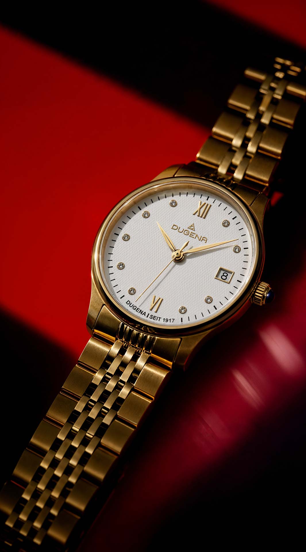 Goldene Dugena-Armbanduhr mit weißem Zifferblatt, römischen Zahlen und Datum auf rotem Hintergrund.