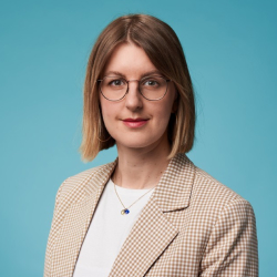 Portrait einer Frau mit Brille, kurzen blonden Haaren, kariertem Blazer und weißem Shirt vor blauem Hintergrund.