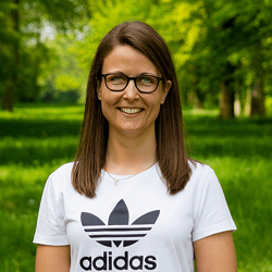 Lächelnde Frau mit braunen Haaren und Brille trägt ein weißes Adidas-T-Shirt im Park.