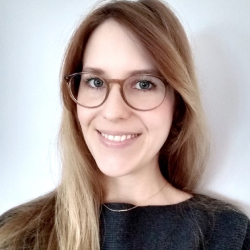 Frau mit langem blonden Haar, Brille und dunklem Oberteil lächelt vor weißem Hintergrund.
