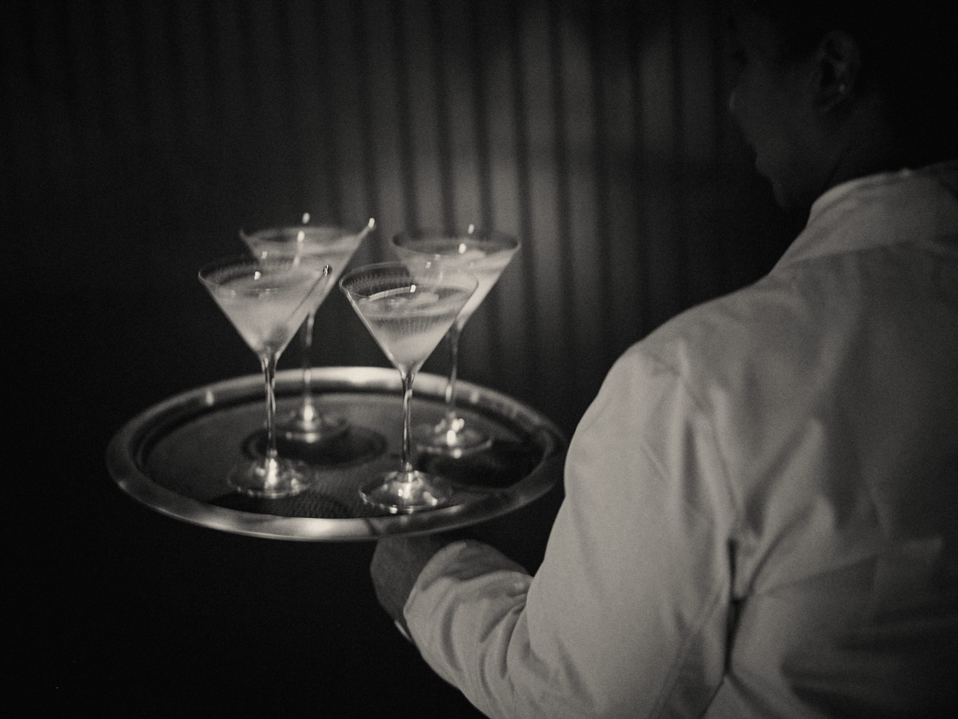 Black and white photo of a server carrying a tray of four martinis through a dark dining room