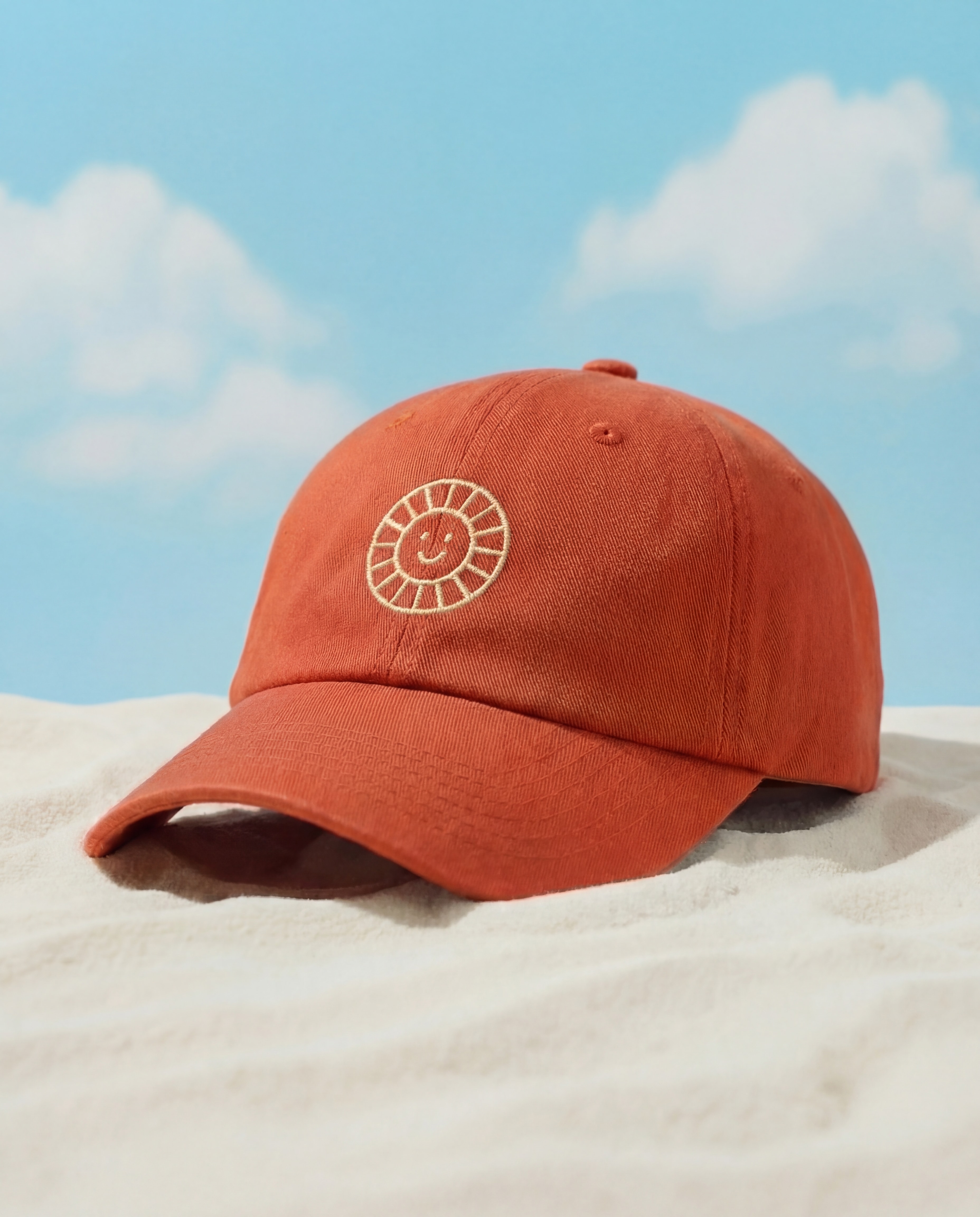 Rust-orange embroidered Daypass cap resting on sand at the beach