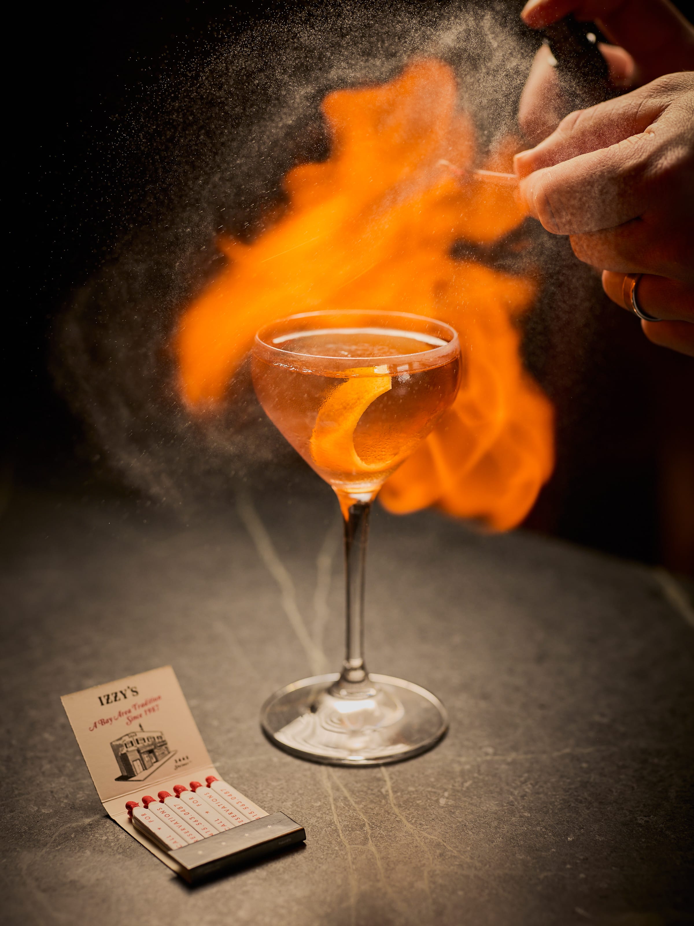 Bartender flaming an orange peel over a cocktail at Izzy's, with Izzy's matchbook in the foreground