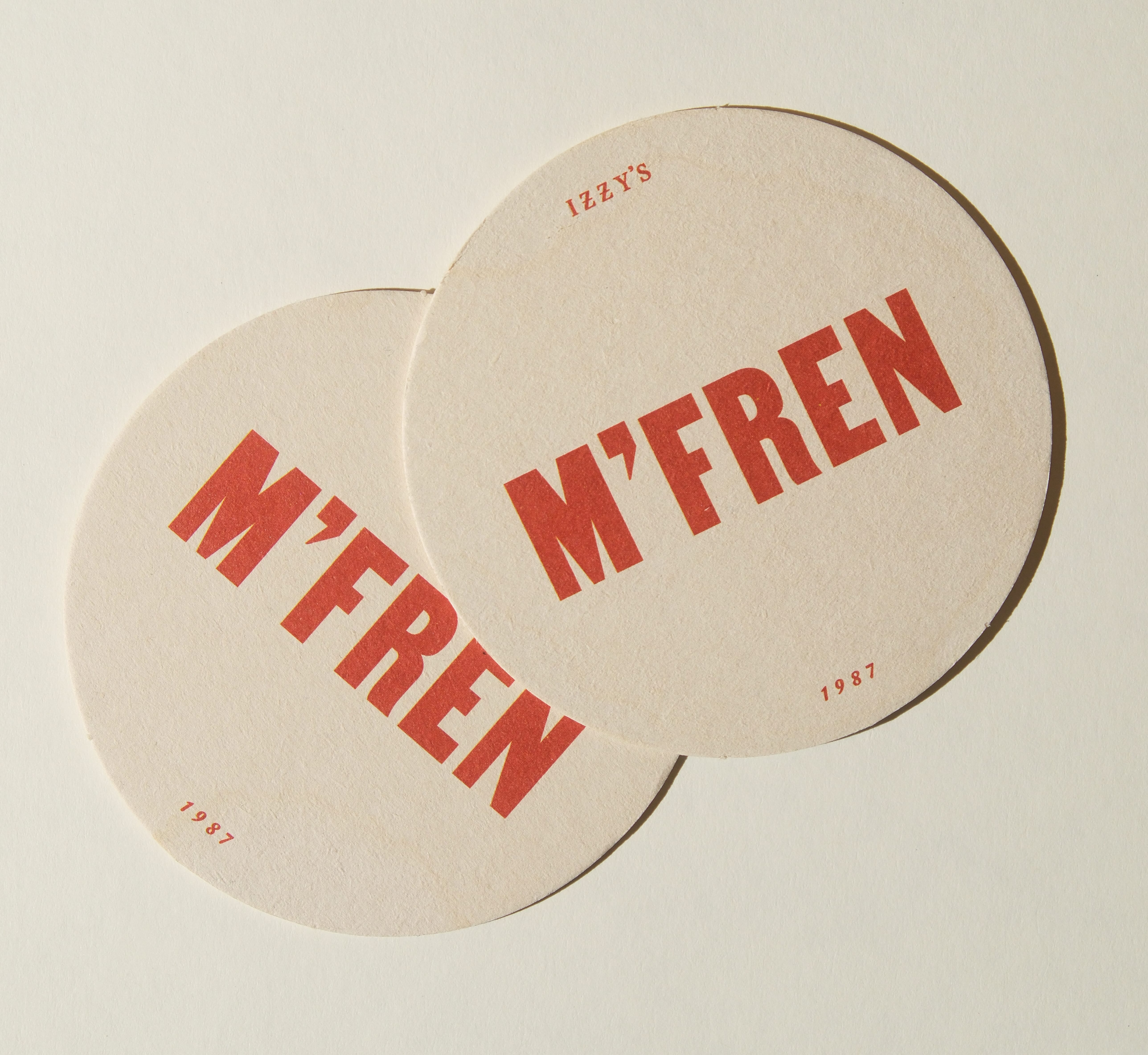 Two round Izzy's bar coasters printed with 'M'FREN' in bold red type and '1987'