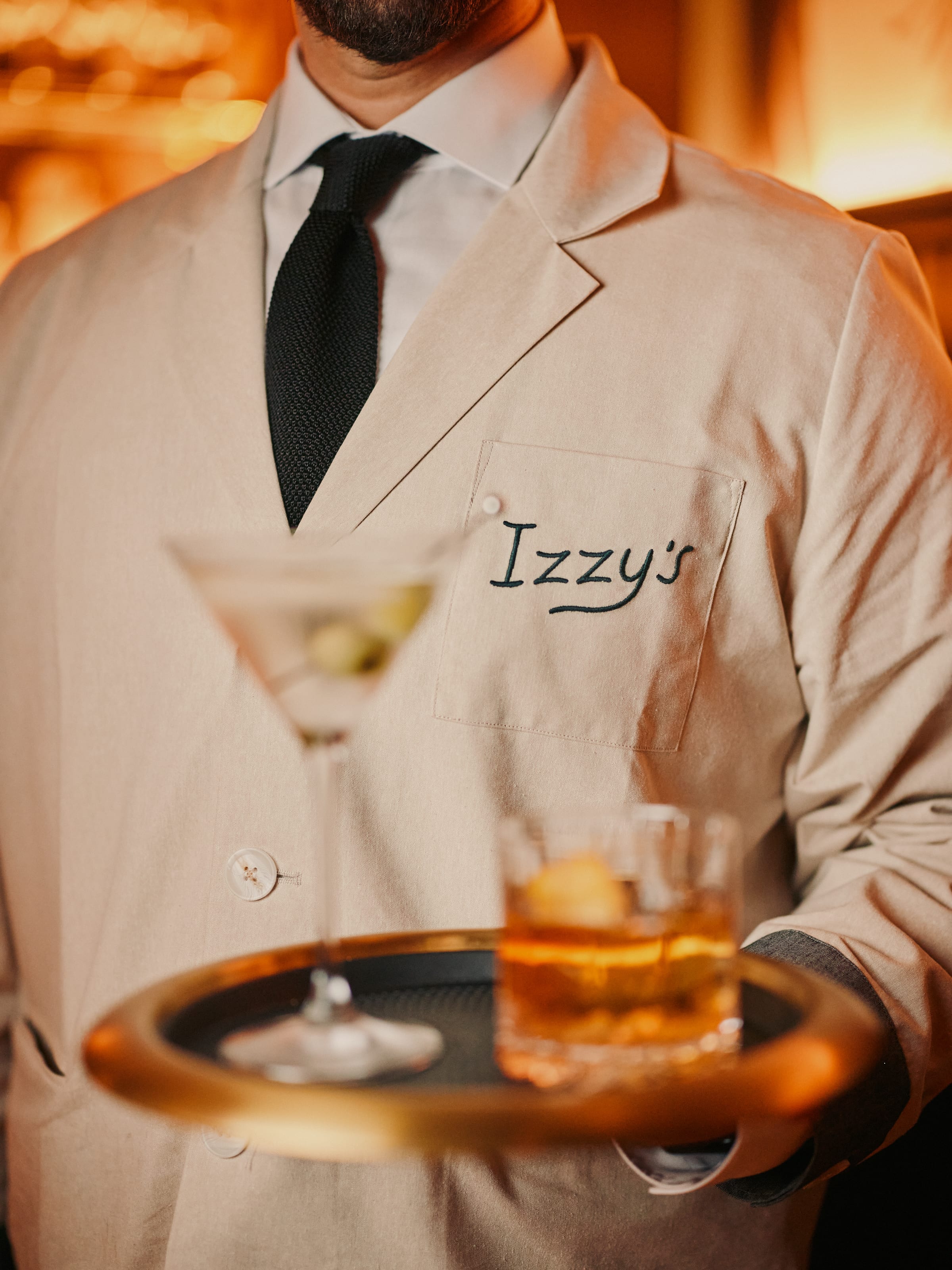 Server in Izzy's embroidered cream jacket carrying a tray with a martini and old fashioned