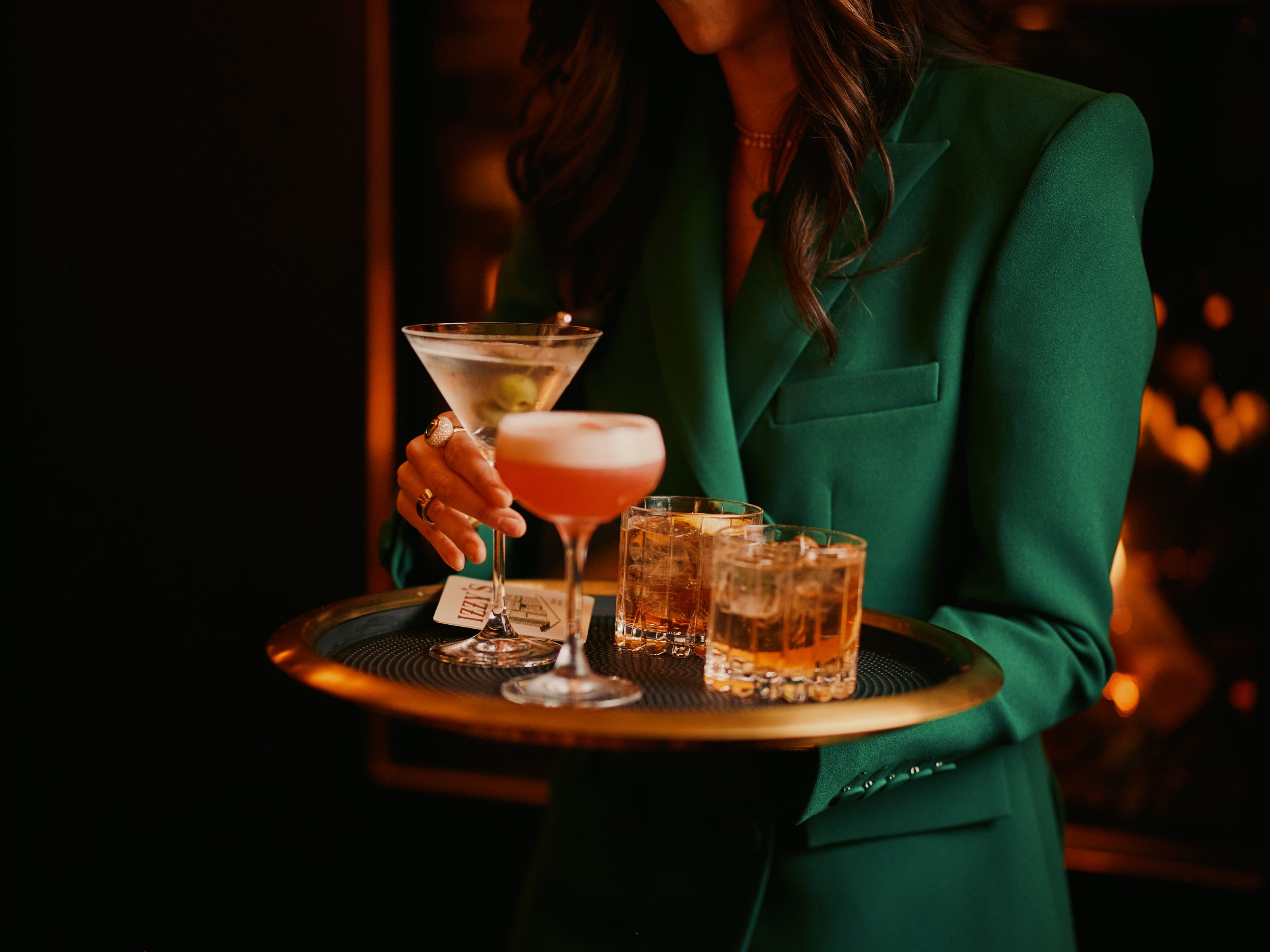 Woman in green blazer carrying a gold tray of cocktails — martini, cosmopolitan, and old fashioneds — in the Izzy's bar