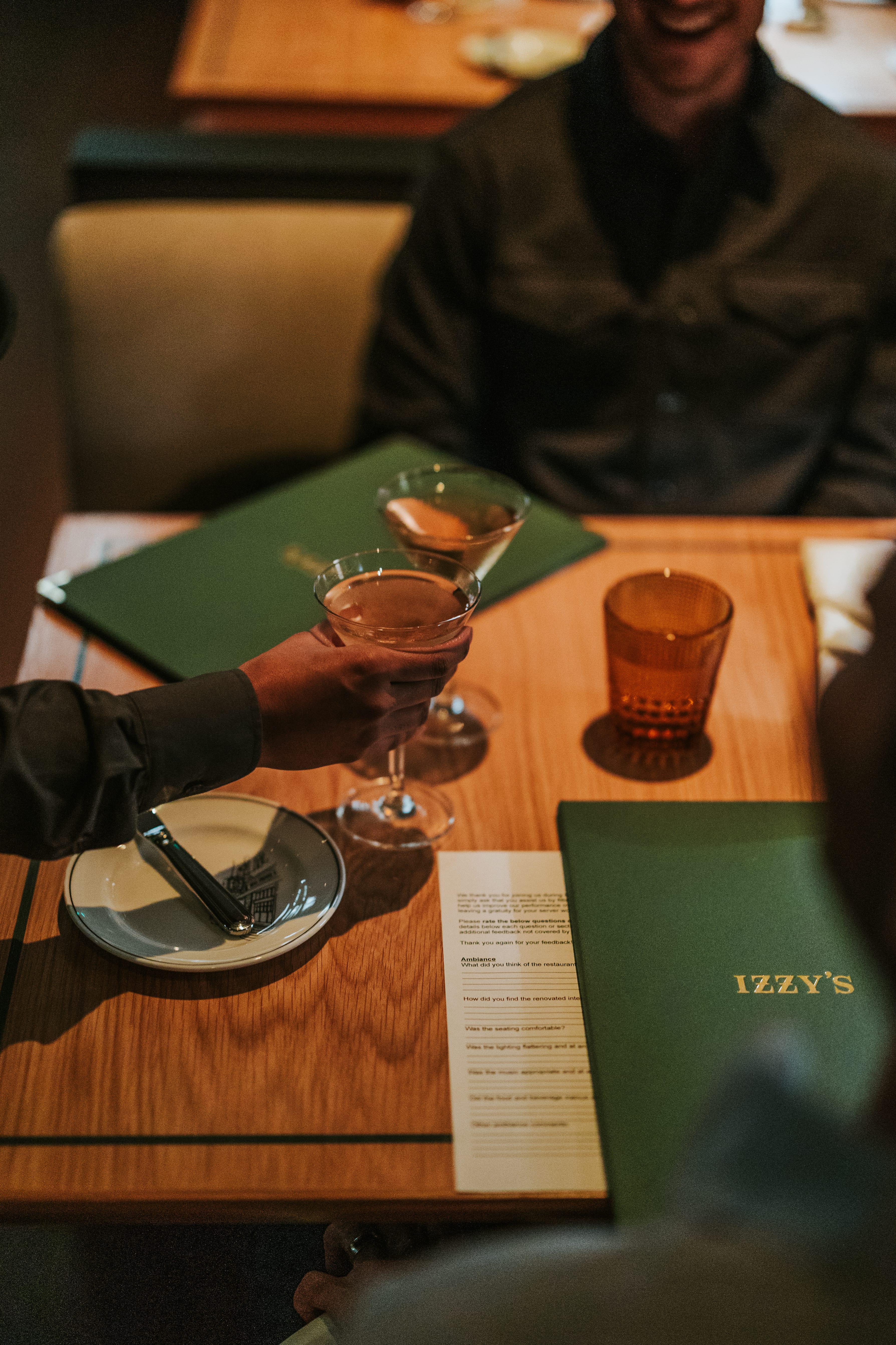 Diner holding a cocktail at a table next to a green Izzy's menu in the candlelit dining room