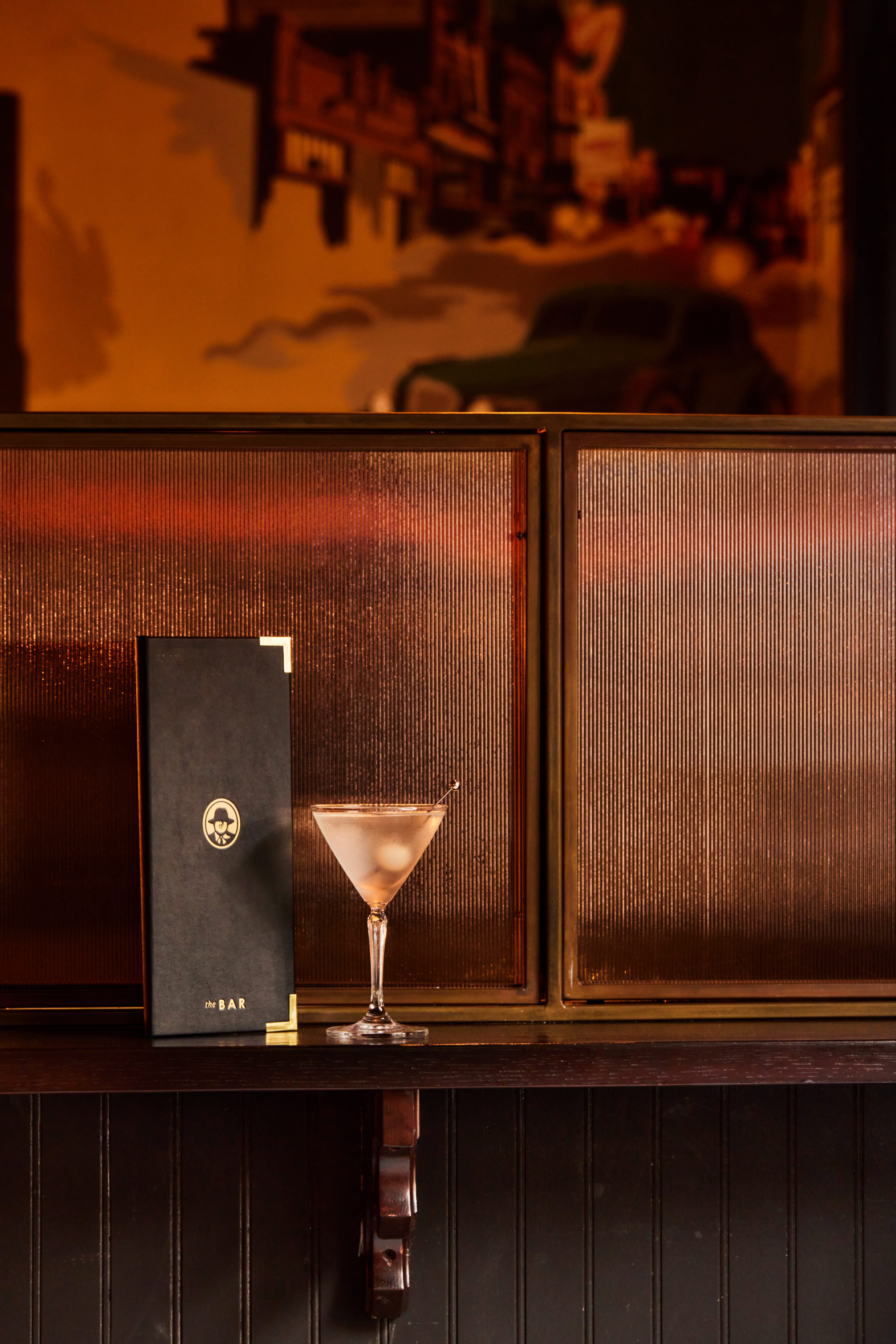 Izzy's bar menu standing next to a cocktail on the bar, amber-lit with a vintage painted mural behind