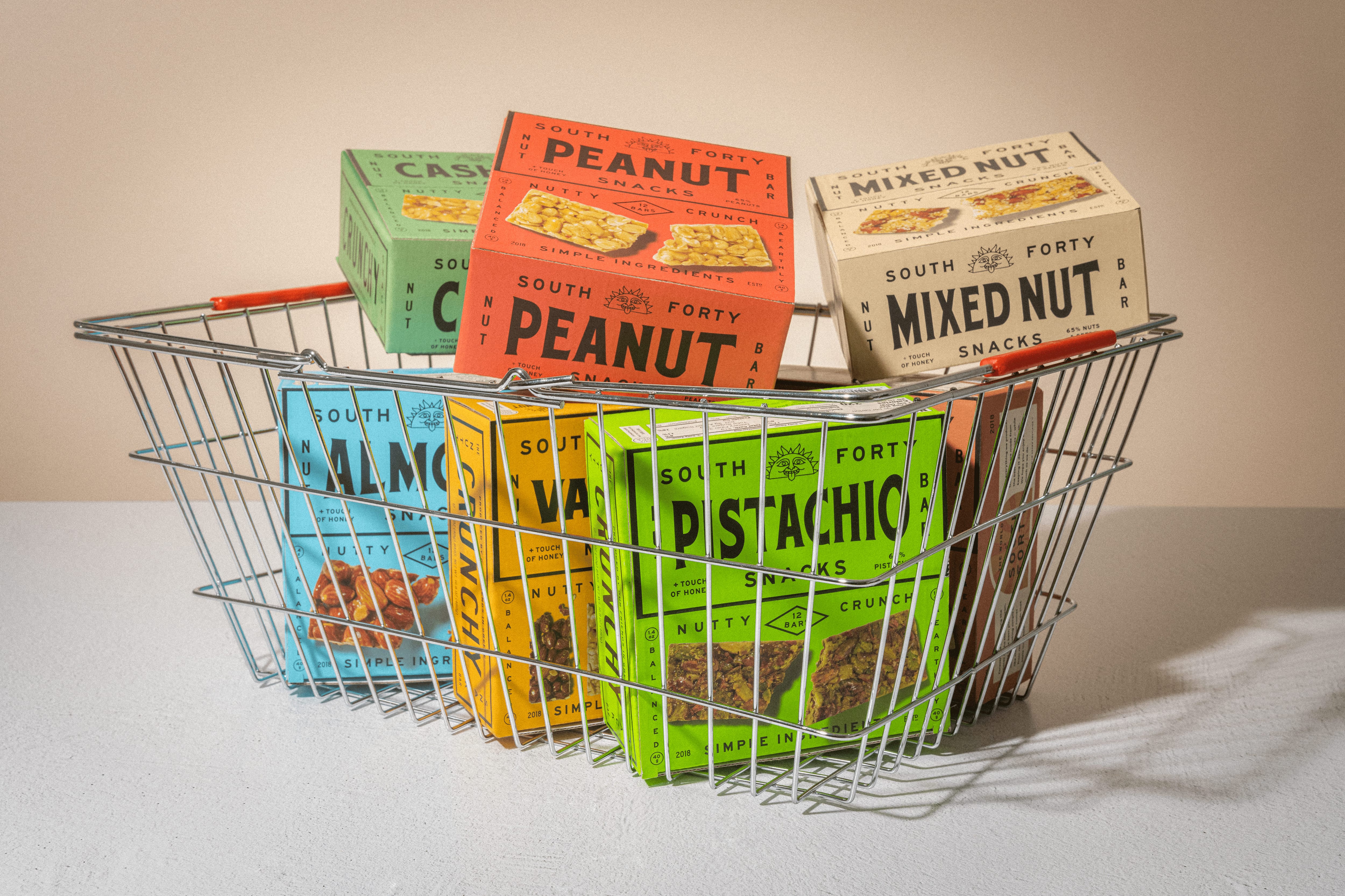 Wire shopping basket filled with all South Forty nut bar flavors — Almond, Peanut, Pistachio, Hazelnut, Cashew, and Mixed Nut
