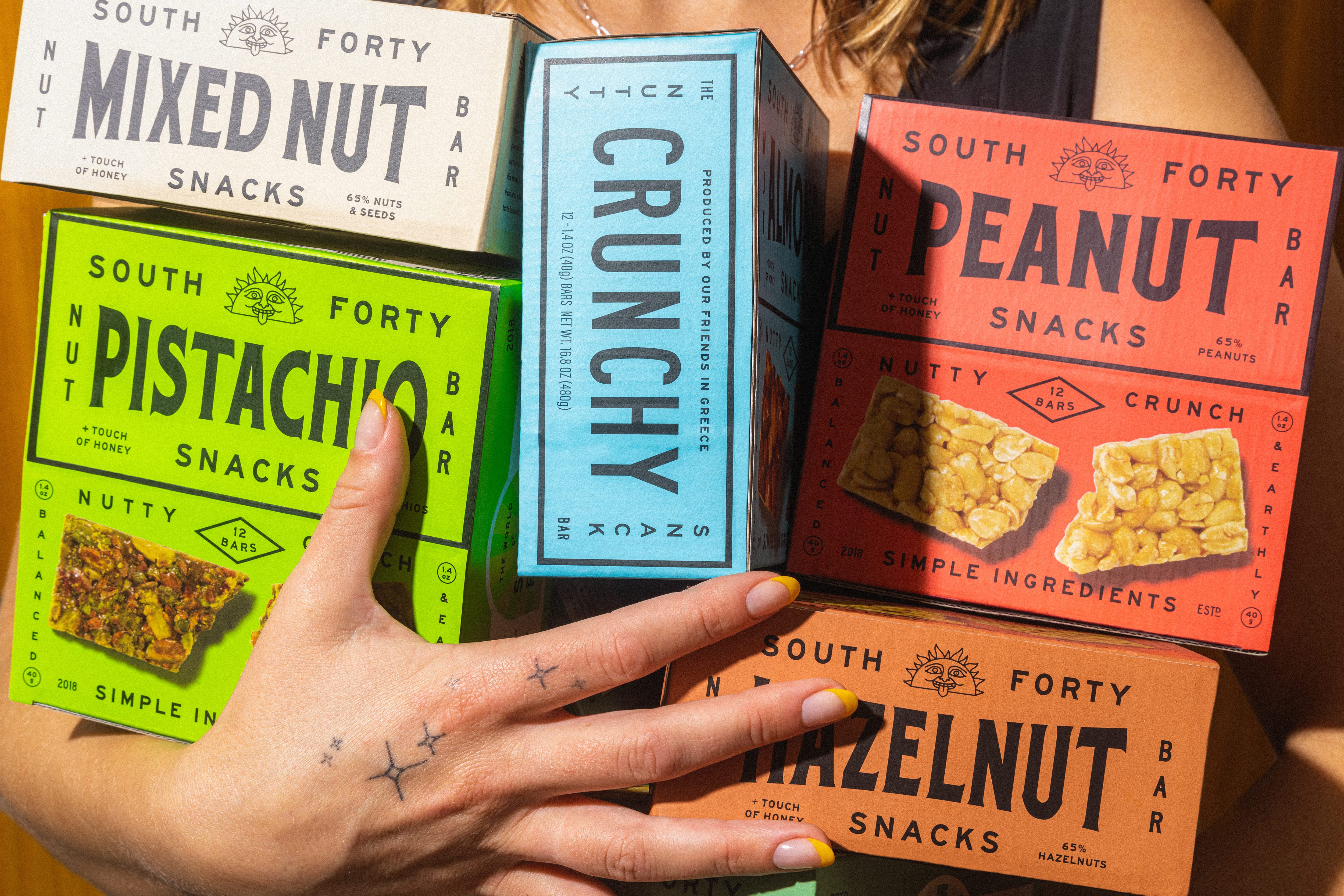 Hands fanning out all South Forty nut bar box flavors — Mixed Nut, Pistachio, Peanut, Hazelnut, and Crunchy varieties