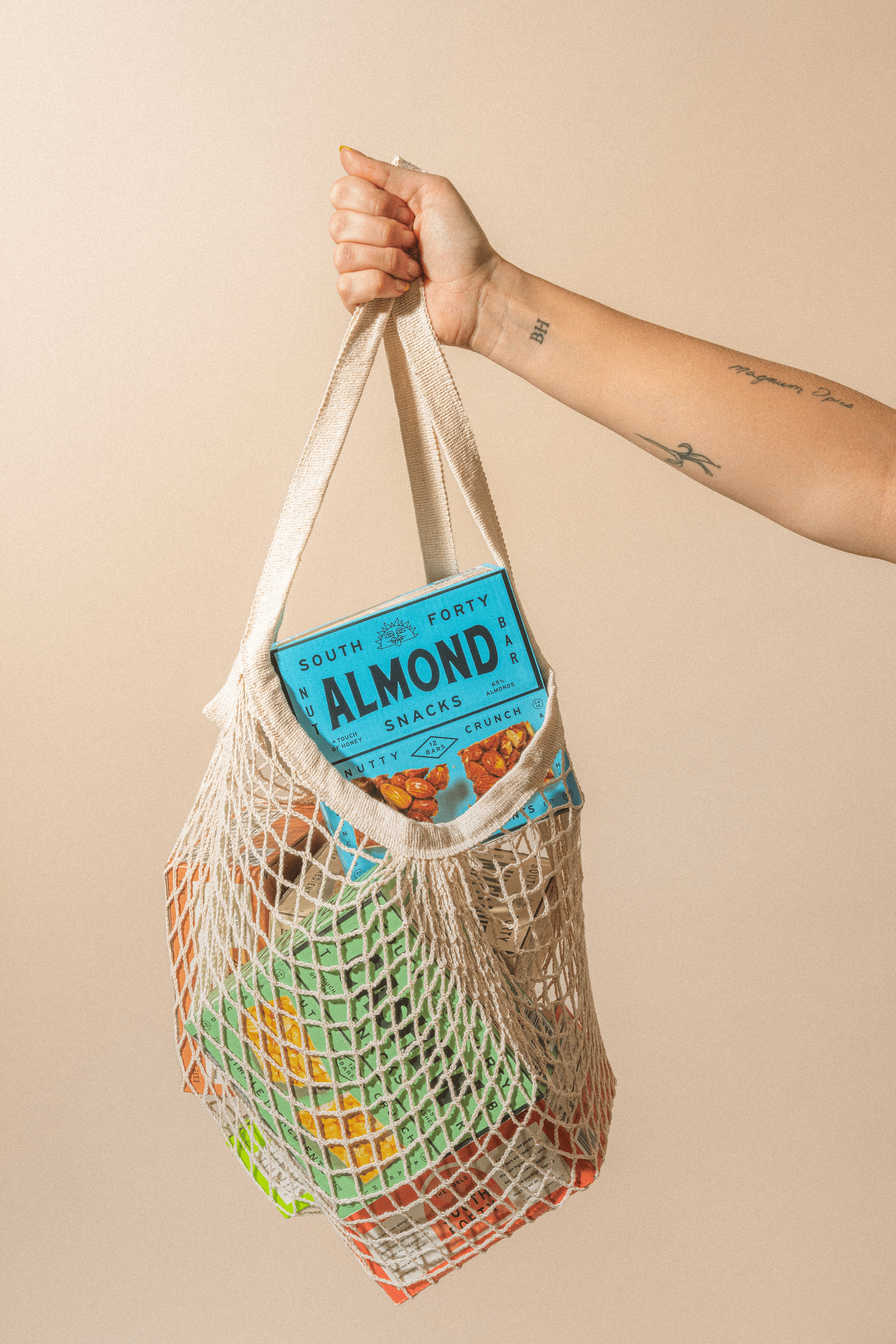 Hand holding a mesh market bag containing South Forty Almond and other nut bar boxes