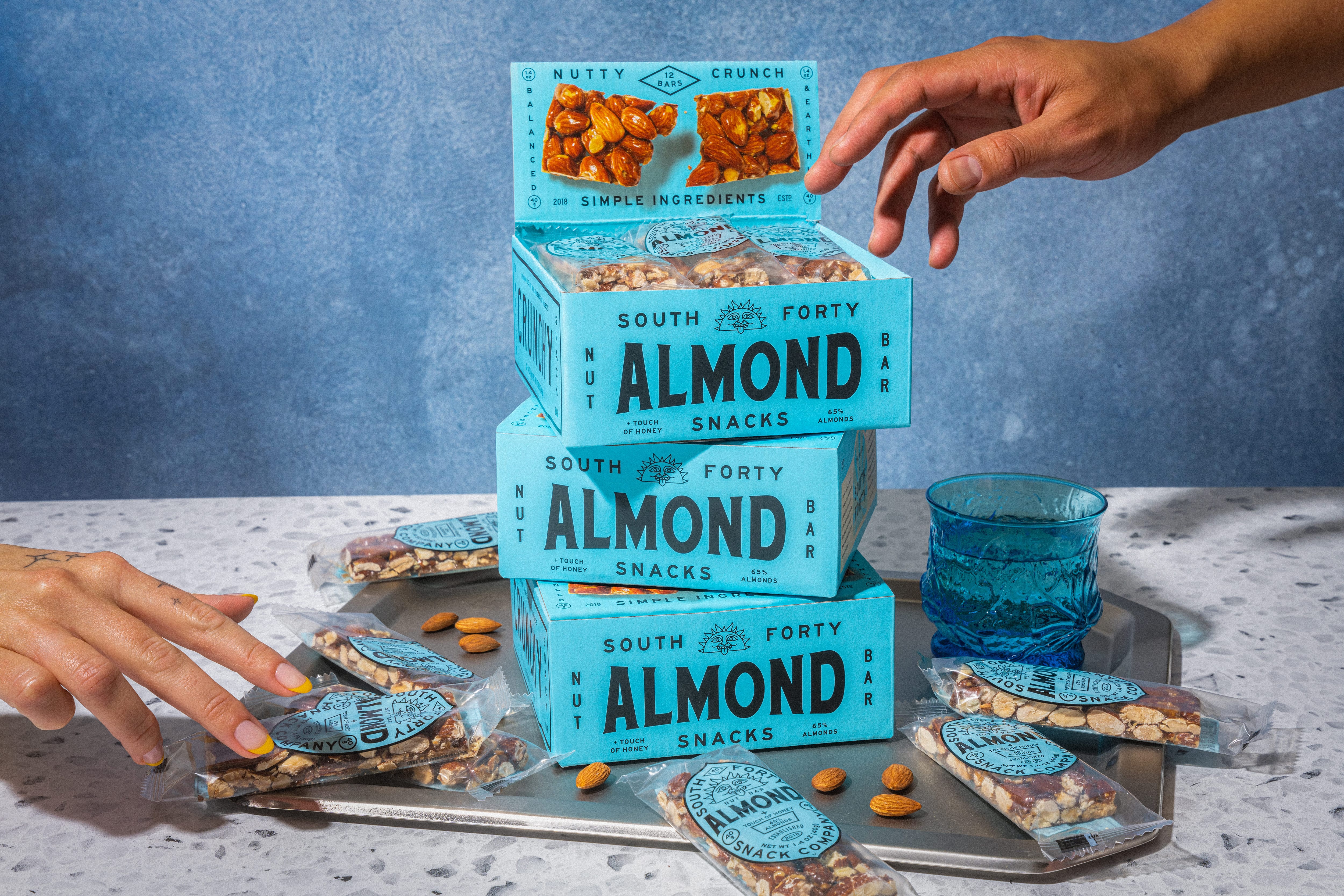 Three stacked South Forty Almond Bar boxes with hands reaching in to grab individual bars from a tray