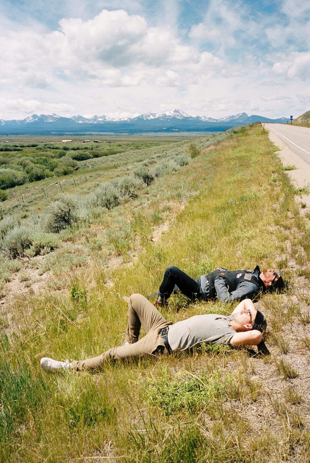 Film photo of two riders lying in tall grass beside a highway with snow-capped mountains in the background
