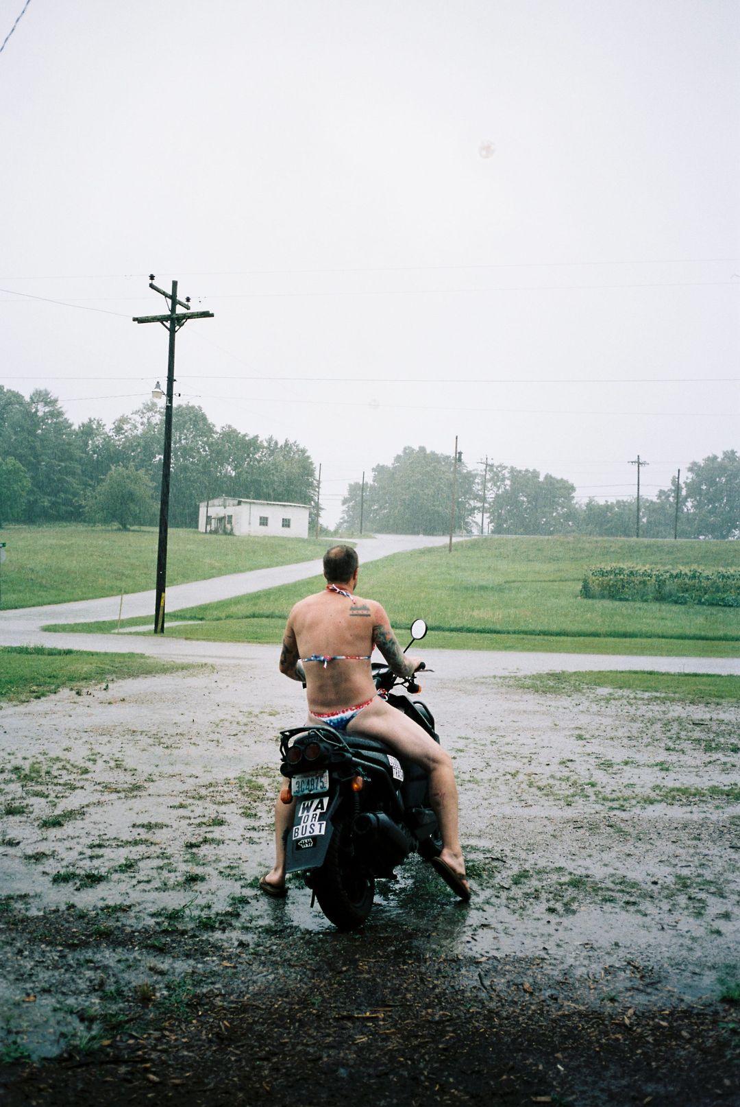 Film photo of a tattooed rider in swim briefs sitting on a motorcycle in a heavy rainstorm on a rural road