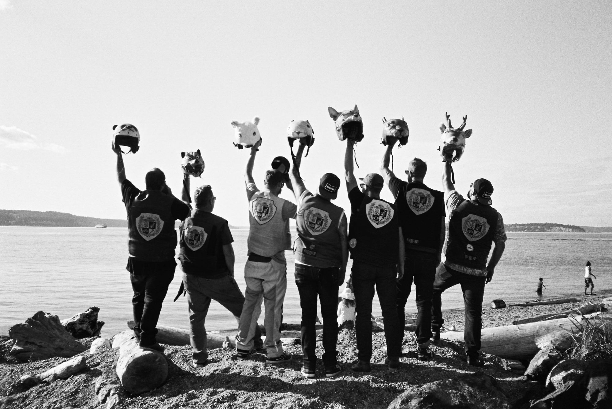 Black and white film photo of Slow Ride Home motorcycle club members holding animal helmet masks aloft on a Pacific Northwest beach