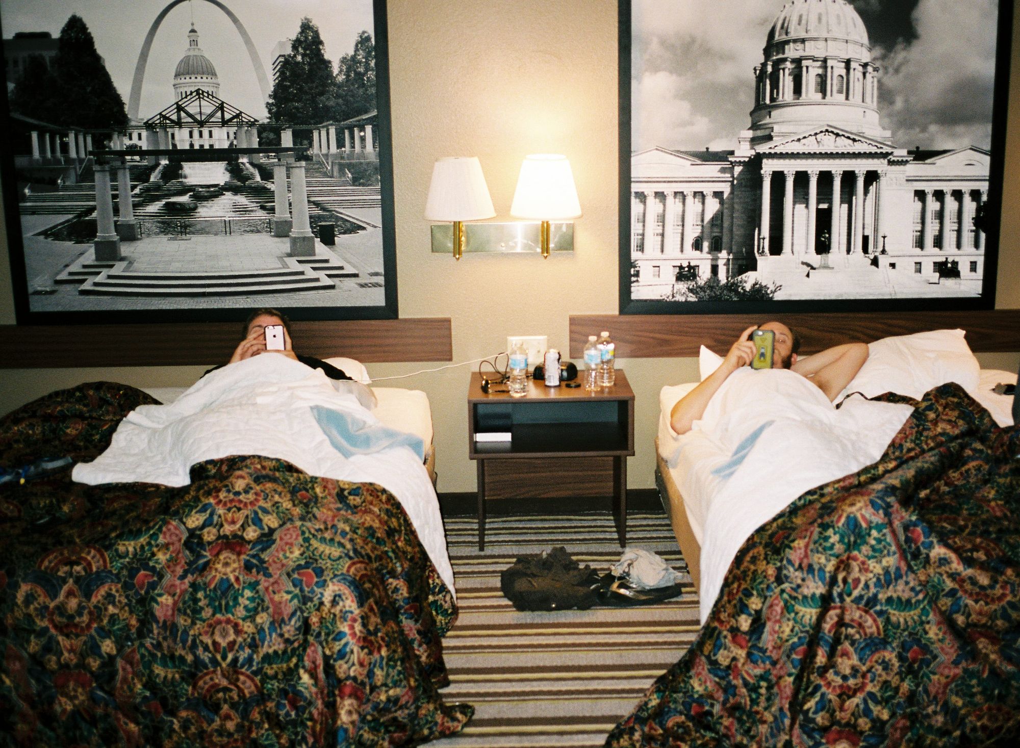 Film photo of two riders resting in motel room double beds with black and white landmark prints on the walls