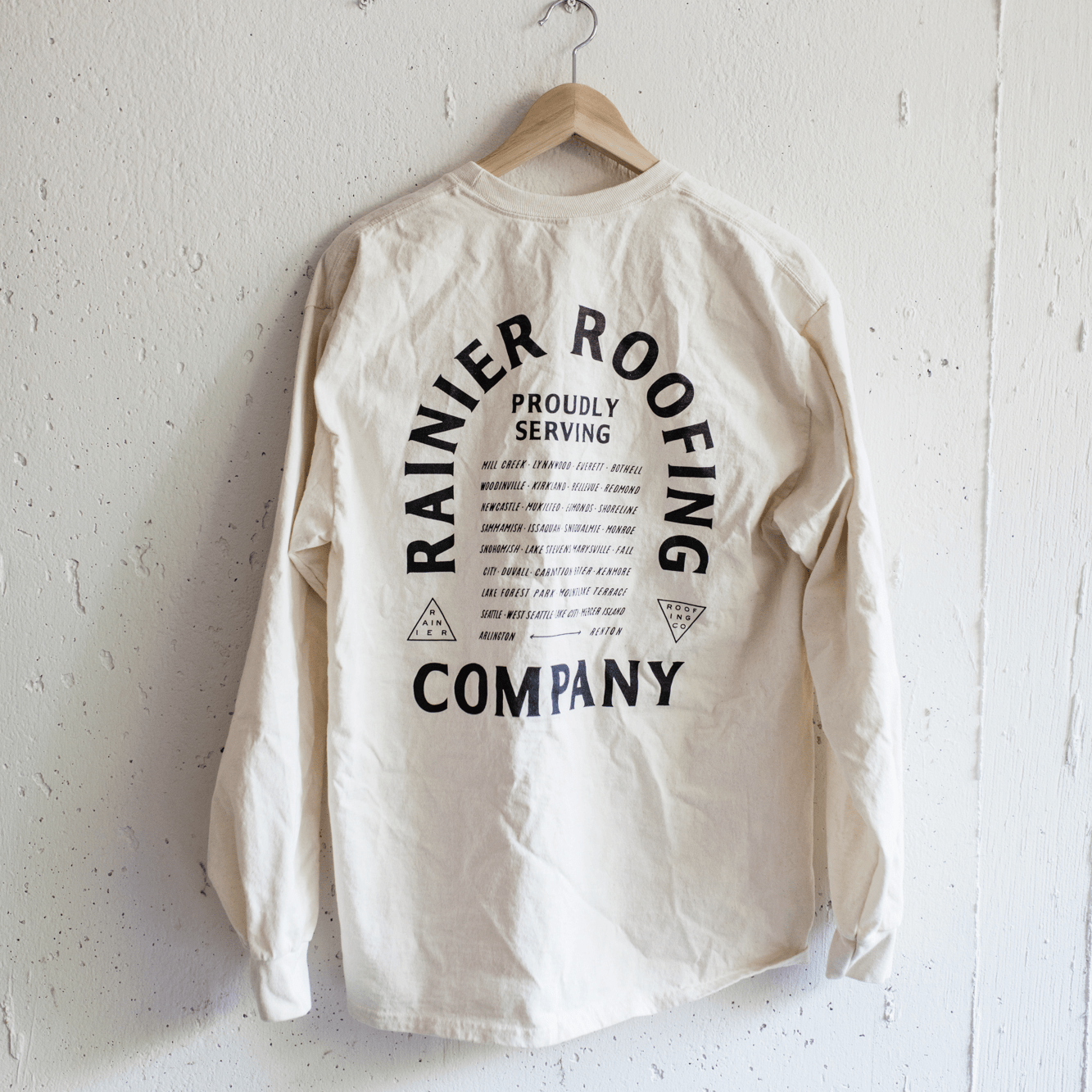 Back of a cream long-sleeve tee with Rainier Roofing Company 'Proudly Serving' graphic and service area cities
