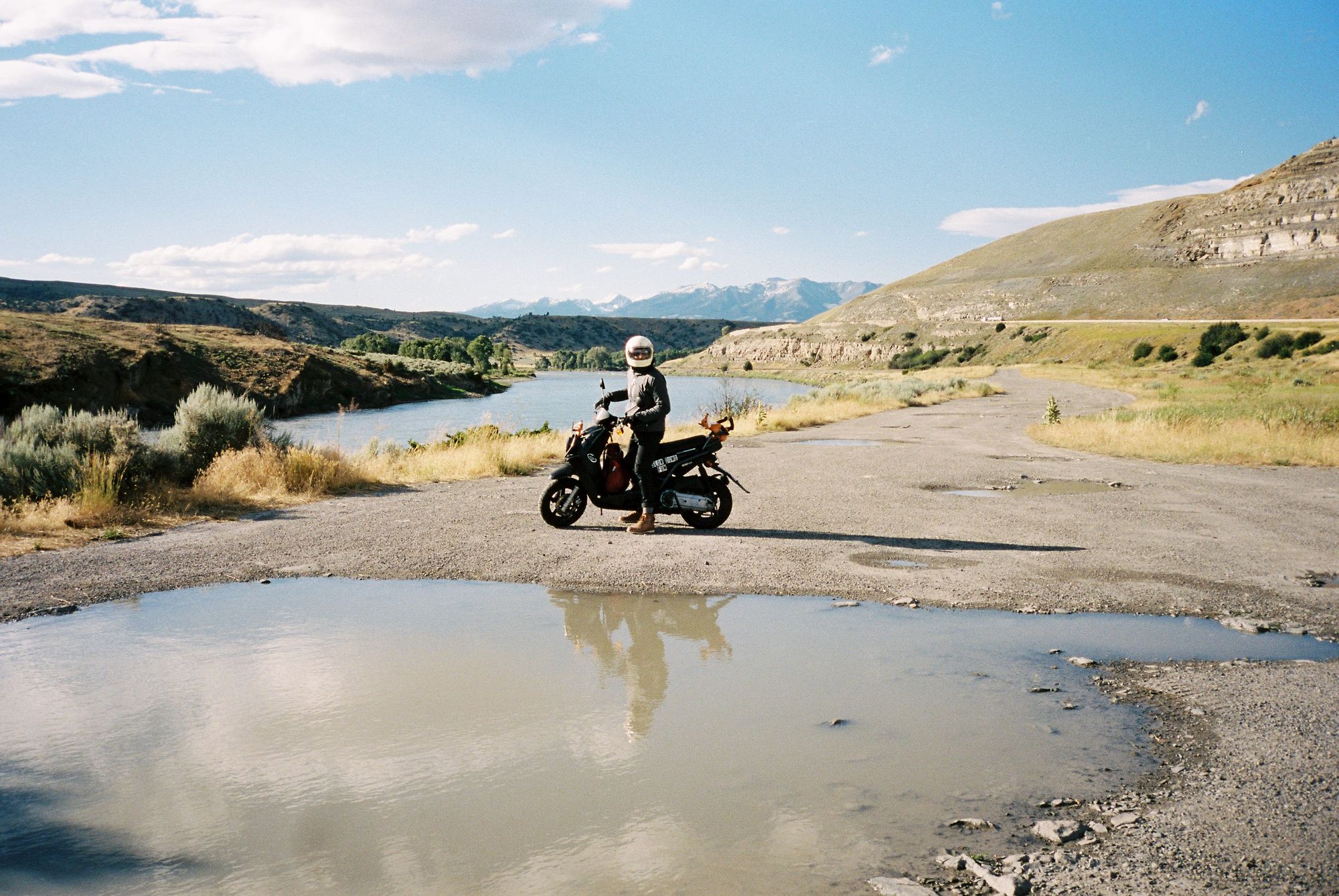 Film photo of a helmeted rider on a loaded scooter reflected in a roadside puddle with mountain river and canyon backdrop