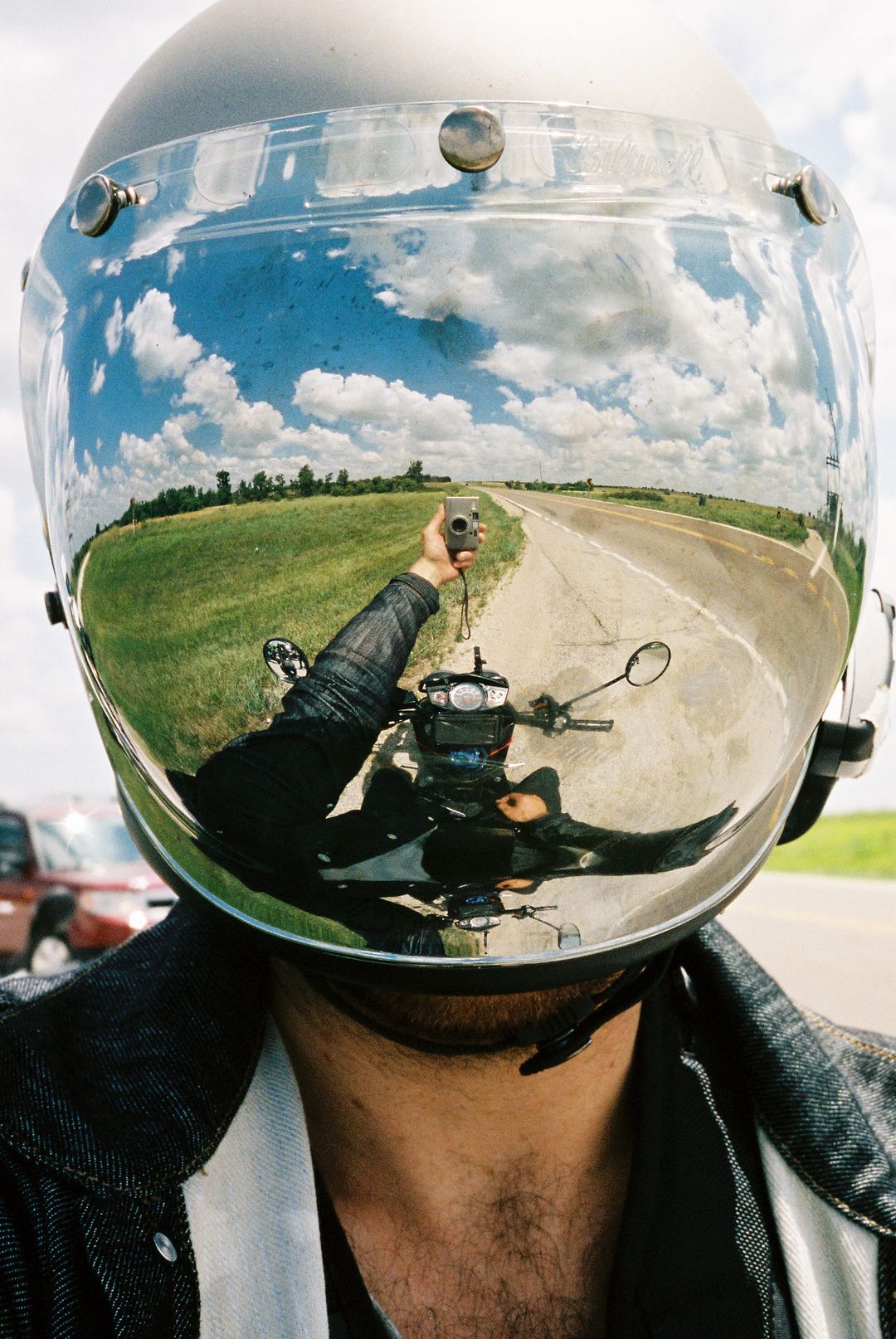Film photo selfie reflected in a chrome motorcycle helmet visor showing the rider and open road under cloudy skies