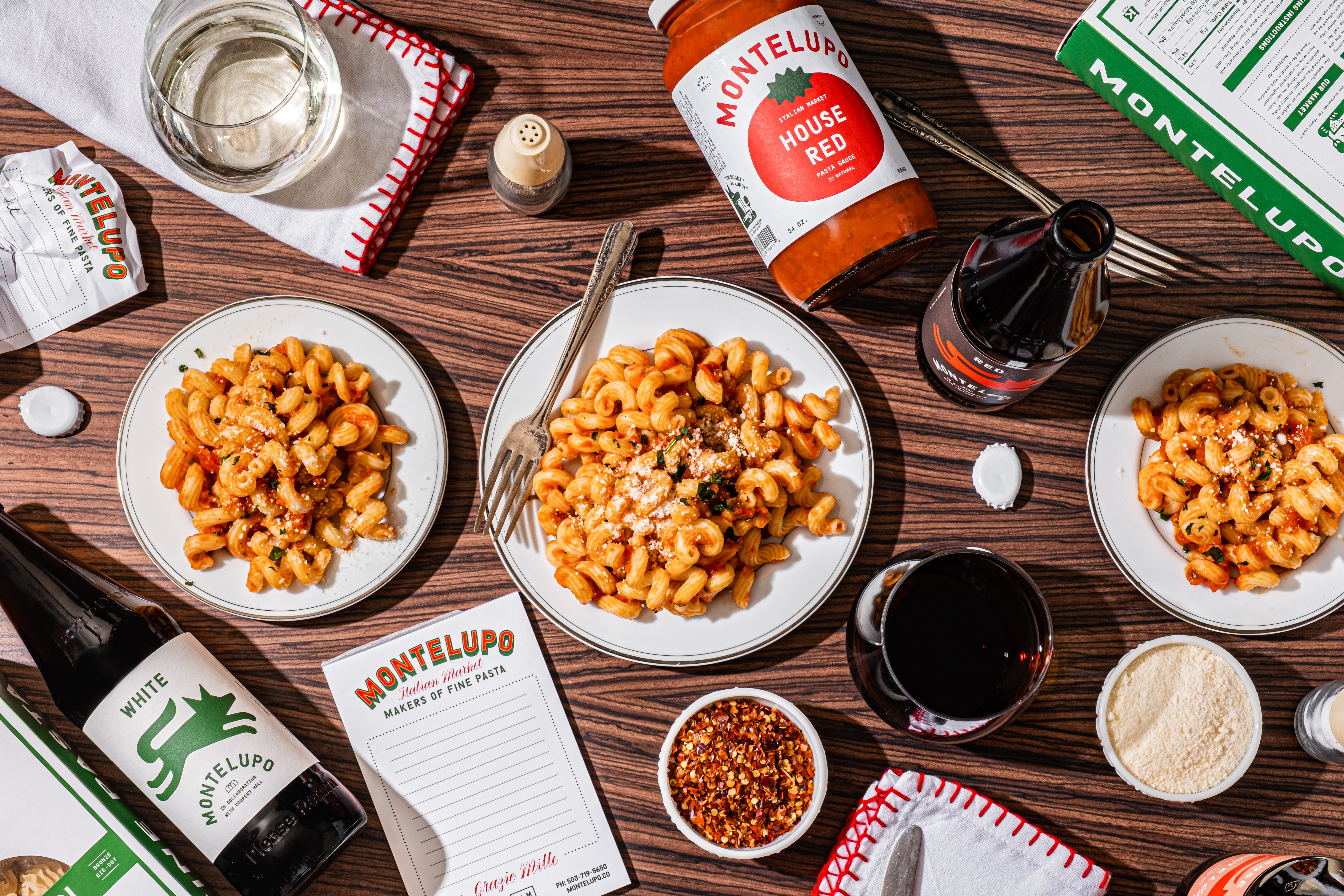 Overhead Montelupo table spread — three plates of pasta, House Red sauce jar, white wine, red wine, and menus
