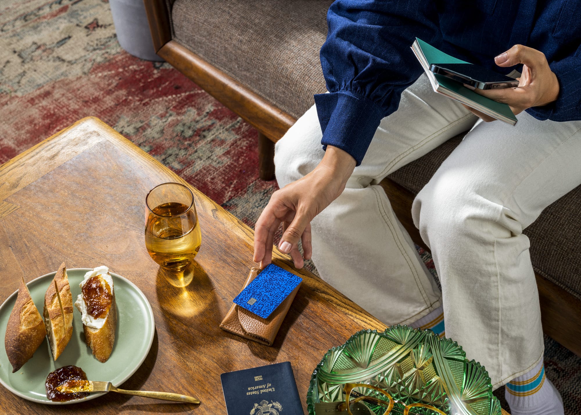 Person on a couch reaching to pick up a blue Venmo card from a woven card holder on a coffee table beside bread and wine