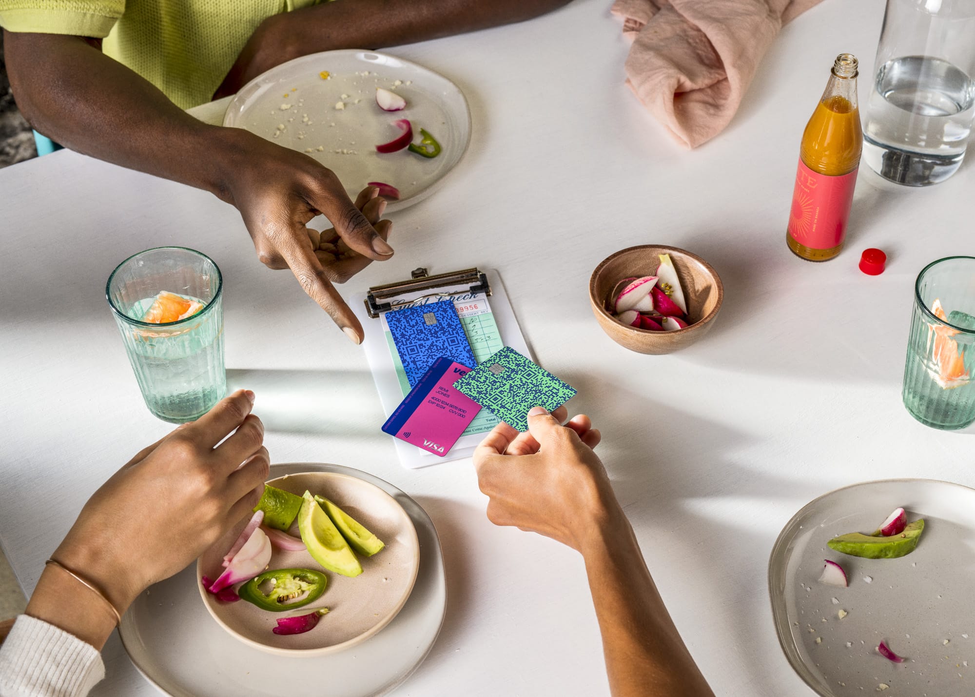 Overhead view of two diners splitting a restaurant bill — two colorful Venmo cards on a clipboard check tray over a white table with avocado plates