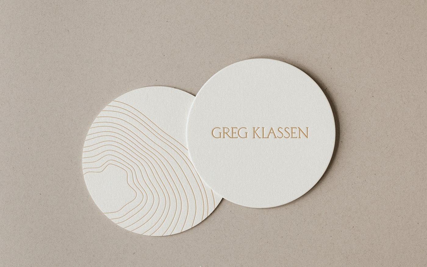 Two overlapping round cream coasters with Greg Klassen gold text and delicate topographic arc line art