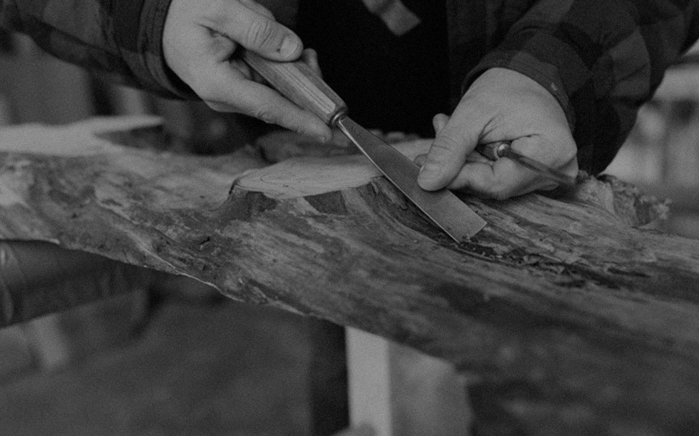 Close-up black and white photo of hands using a chisel to carve a wood slab on a workshop workbench