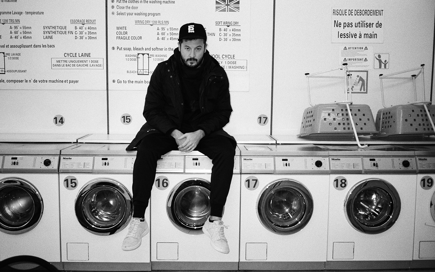 Black and white photo of SYML artist in all-black sitting on top of washing machines in a French laundromat