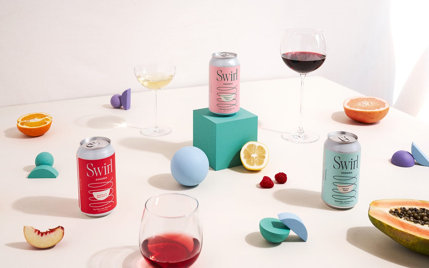 Product lifestyle scene with three Swirl sangria cans surrounded by citrus, papaya, raspberries, wine glasses, and geometric foam shapes on a white surface