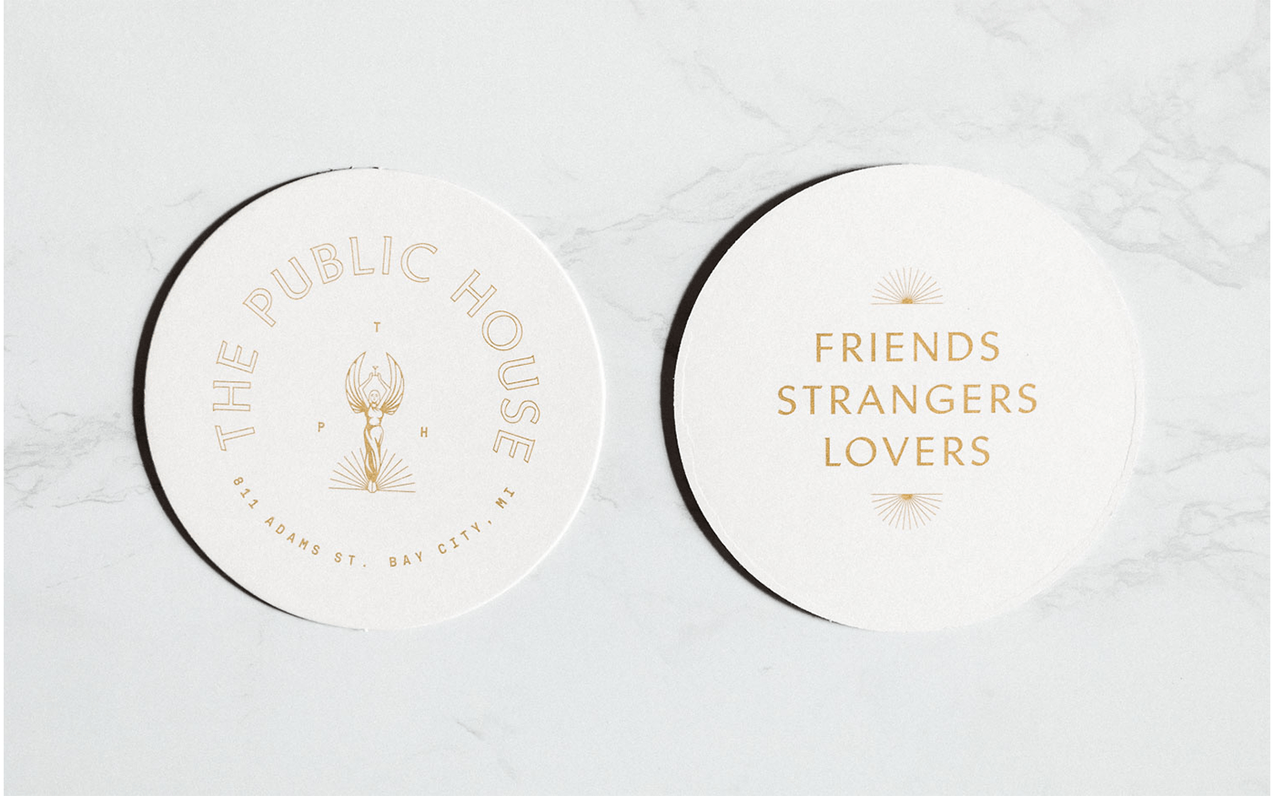 Two round white coasters on marble — left with The Public House goddess logo, right with Friends Strangers Lovers text