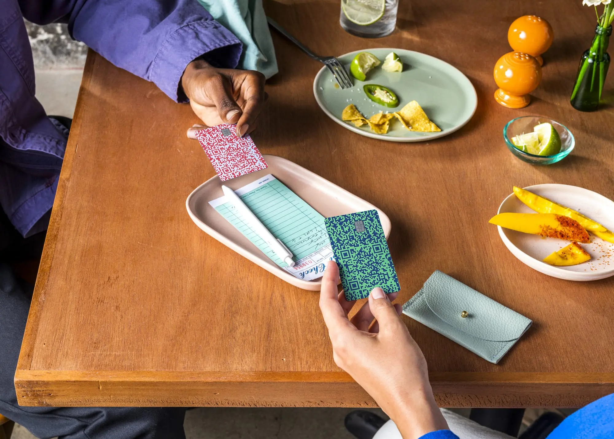 Two diners exchanging colorful Venmo cards over a restaurant check tray with cocktails and tropical food on a wooden table