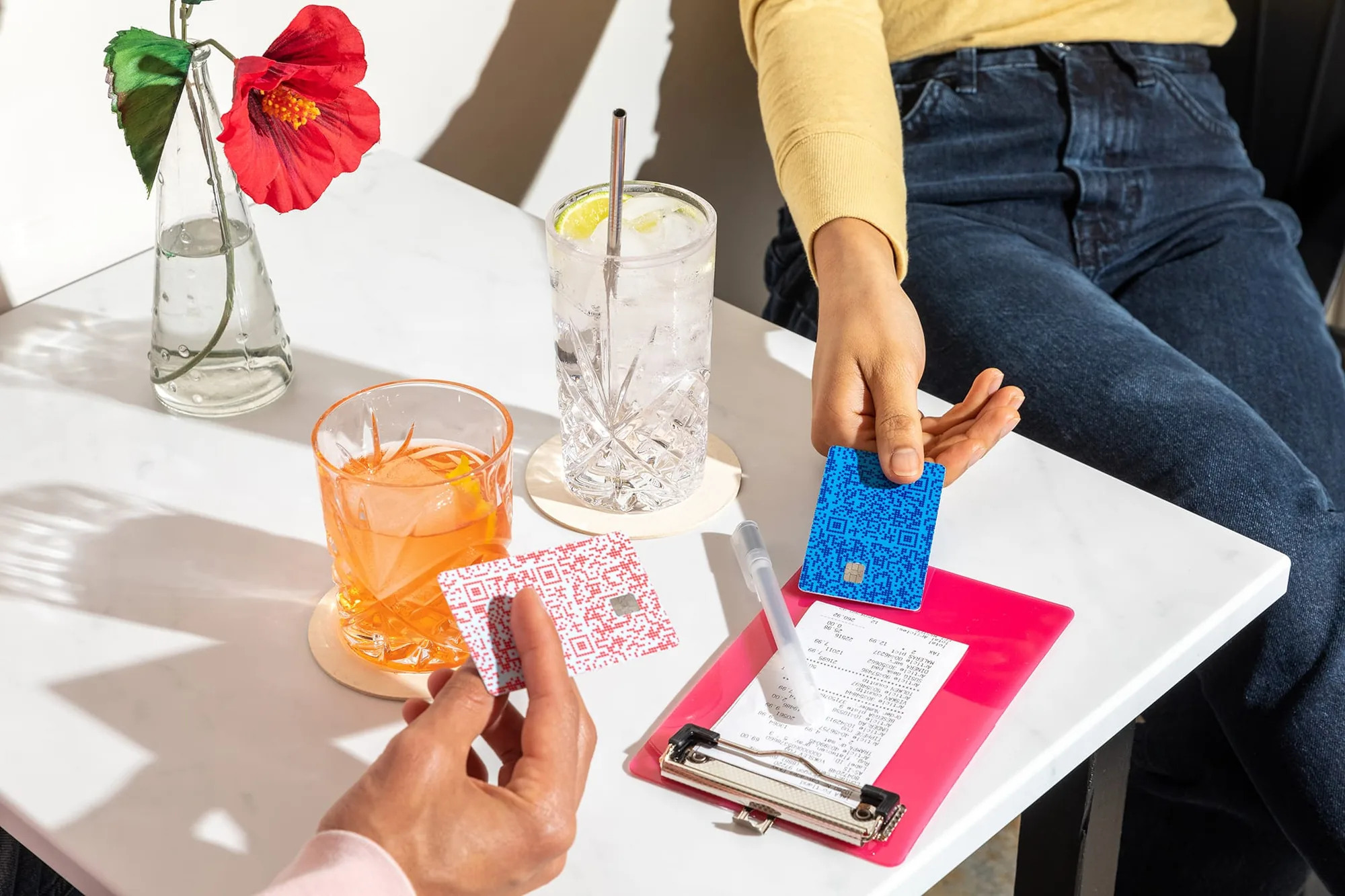 Two people splitting a bar tab — two colorful Venmo cards on a pink clipboard bill tray with cocktails and a red hibiscus flower vase