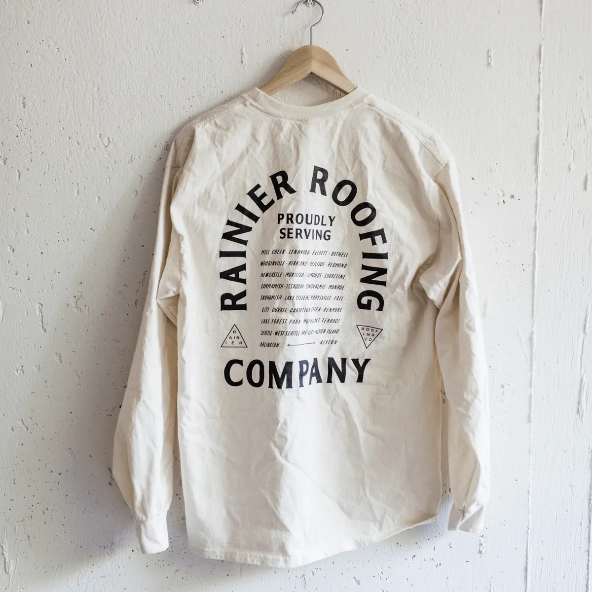 Back of a cream long-sleeve tee with Rainier Roofing Company 'Proudly Serving' graphic and service area cities