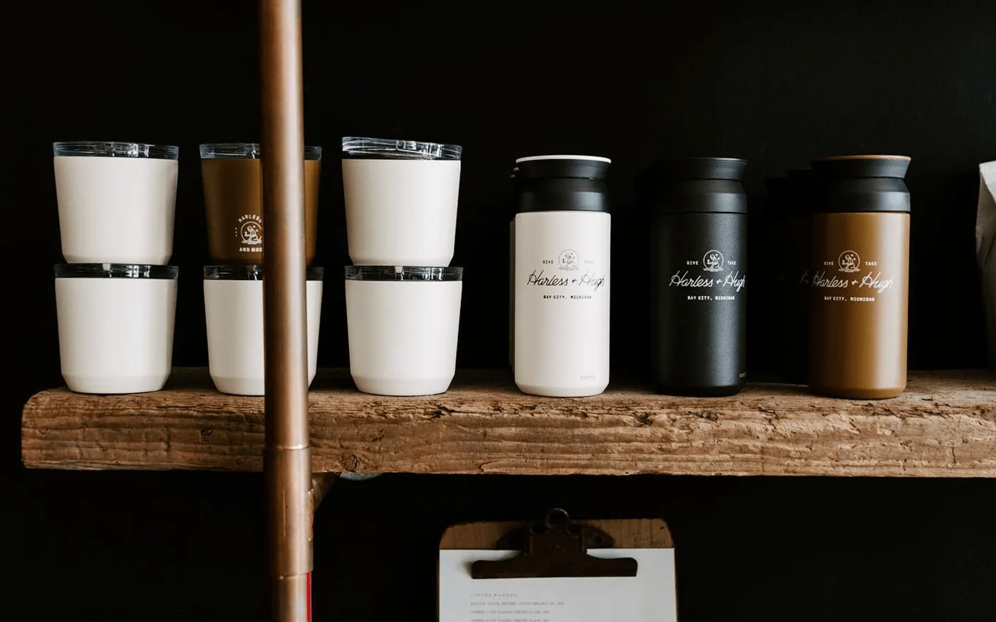 Row of Harless and Hugh branded thermos cups and travel mugs in white, black, and brown on a rustic wooden shelf
