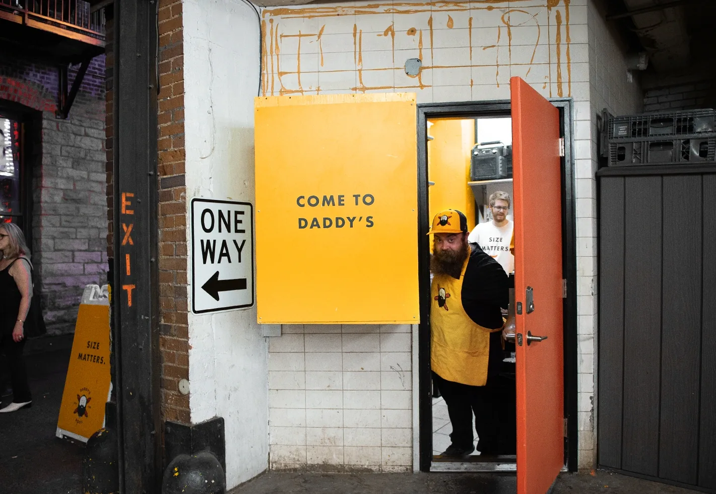Daddy's Dogs employee in yellow uniform and apron holding a door open under a Come to Daddy's yellow sign at a brick building entry