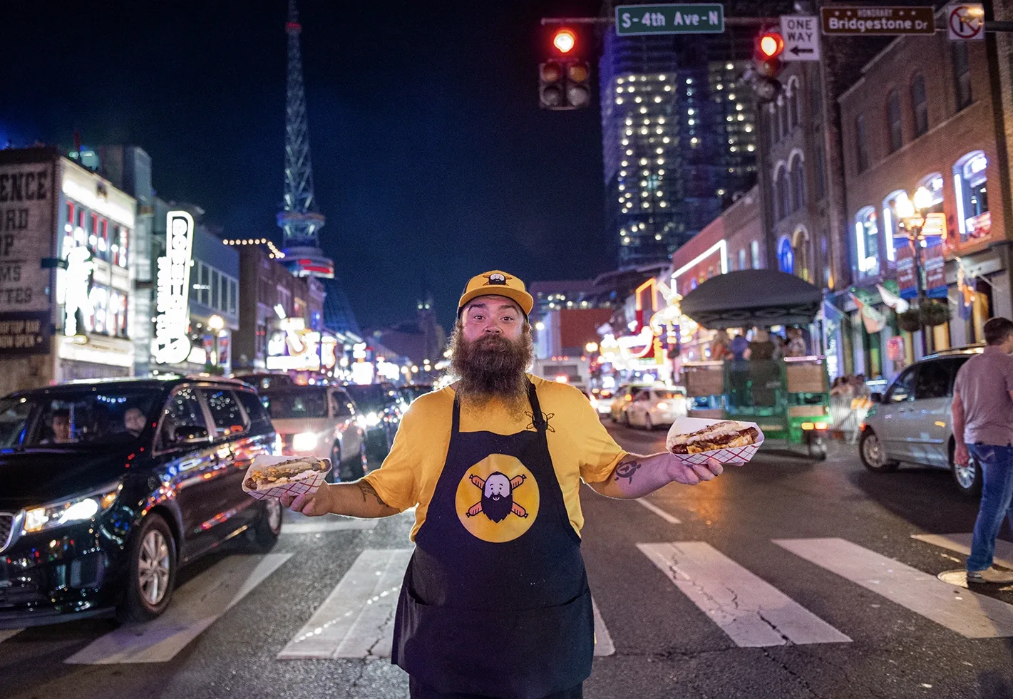 Bearded man in Daddy's Dogs yellow shirt and apron holding two hot dogs on a busy Nashville street at night