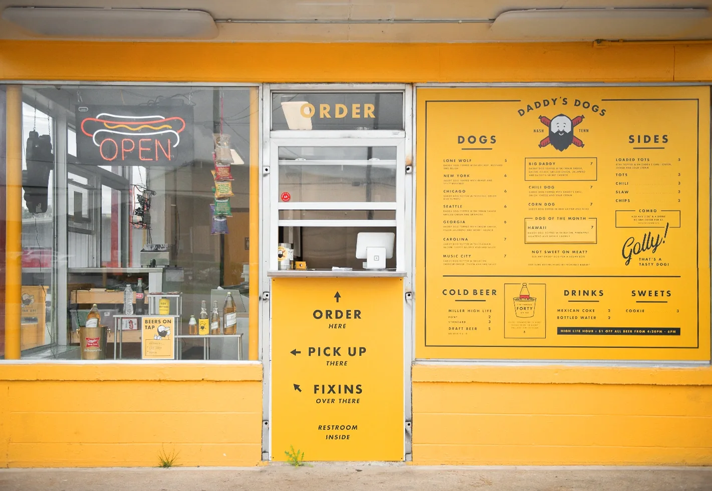 Yellow Daddy's Dogs order window with full menu board and Order, Pick Up, Fixins directional signage on the building exterior