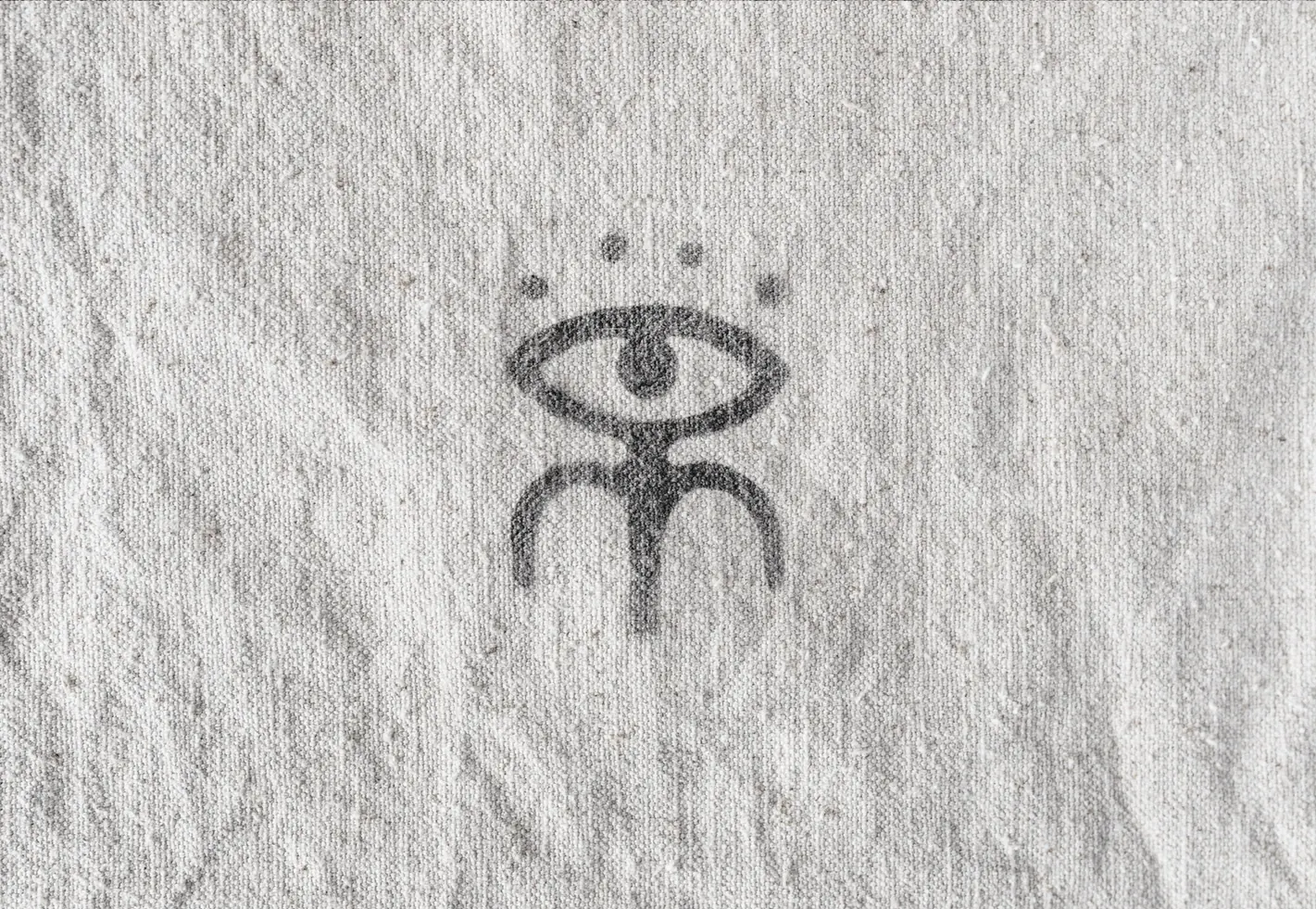 De Melo eye and figure icon stamp pressed in gray on wrinkled linen fabric