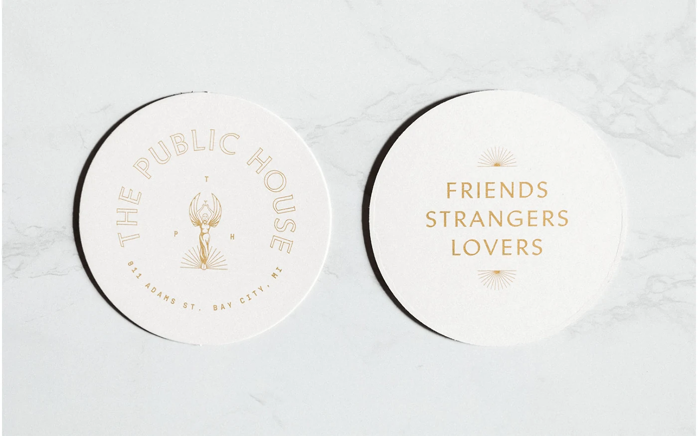 Two round white coasters on marble — left with The Public House goddess logo, right with Friends Strangers Lovers text