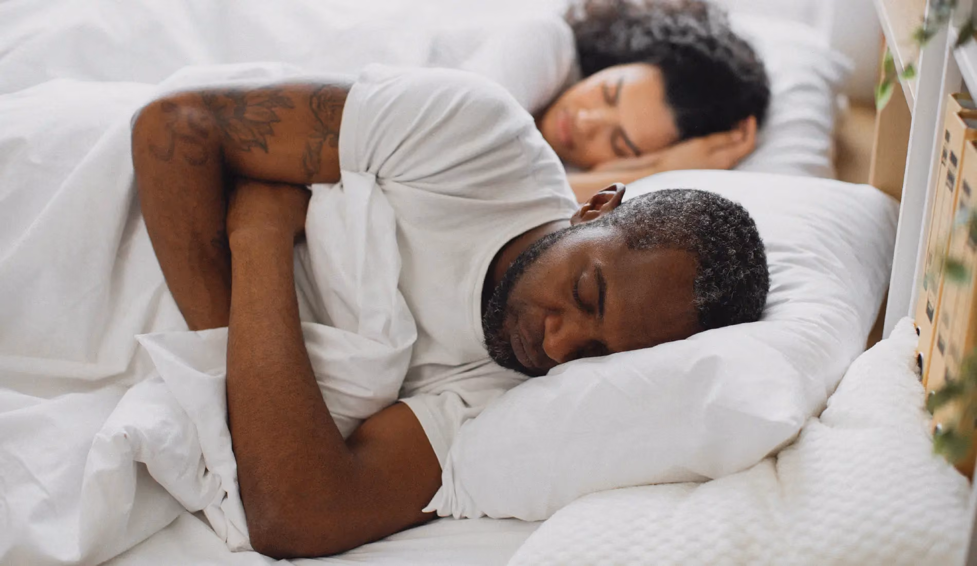 Un couple (homme noir, femme blanche) est couché dans un lit. L'homme tourne le dos à sa femme car elle ronfle.