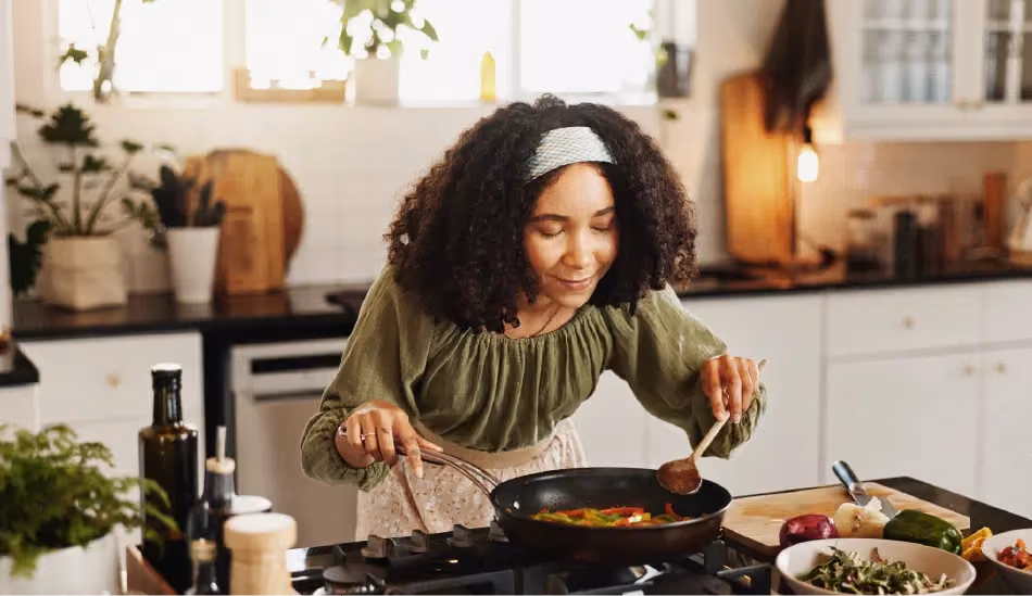 Une jeune dame en train de cuisiner. Elle est penchée vers l'avant, les mains tenant des ustensiles de cuisine posés dans une casserole. Elle sourit.