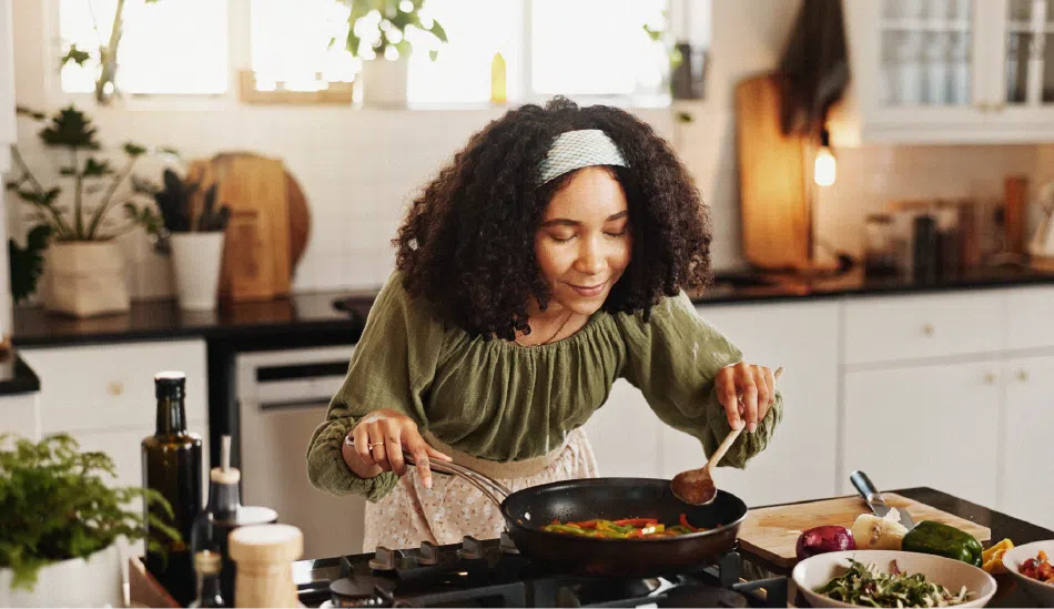 Une jeune dame en train de cuisiner. Elle est penchée vers l'avant, les mains tenant des ustensiles de cuisine posés dans une casserole. Elle sourit.