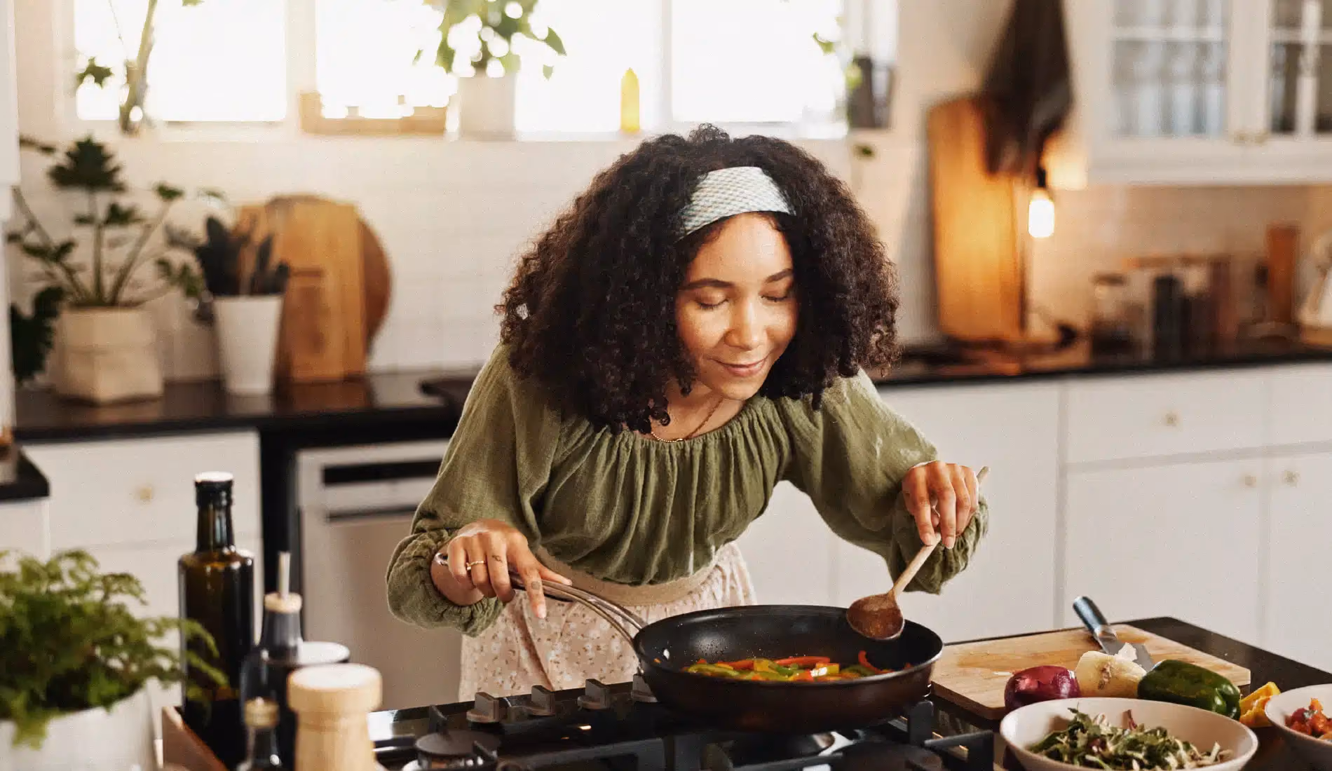 Une jeune dame en train de cuisiner. Elle est penchée vers l'avant, les mains tenant des ustensiles de cuisine posés dans une casserole. Elle sourit.