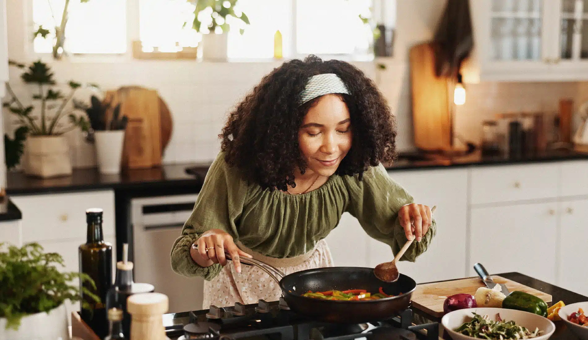 Une jeune dame en train de cuisiner. Elle est penchée vers l'avant, les mains tenant des ustensiles de cuisine posés dans une casserole. Elle sourit.