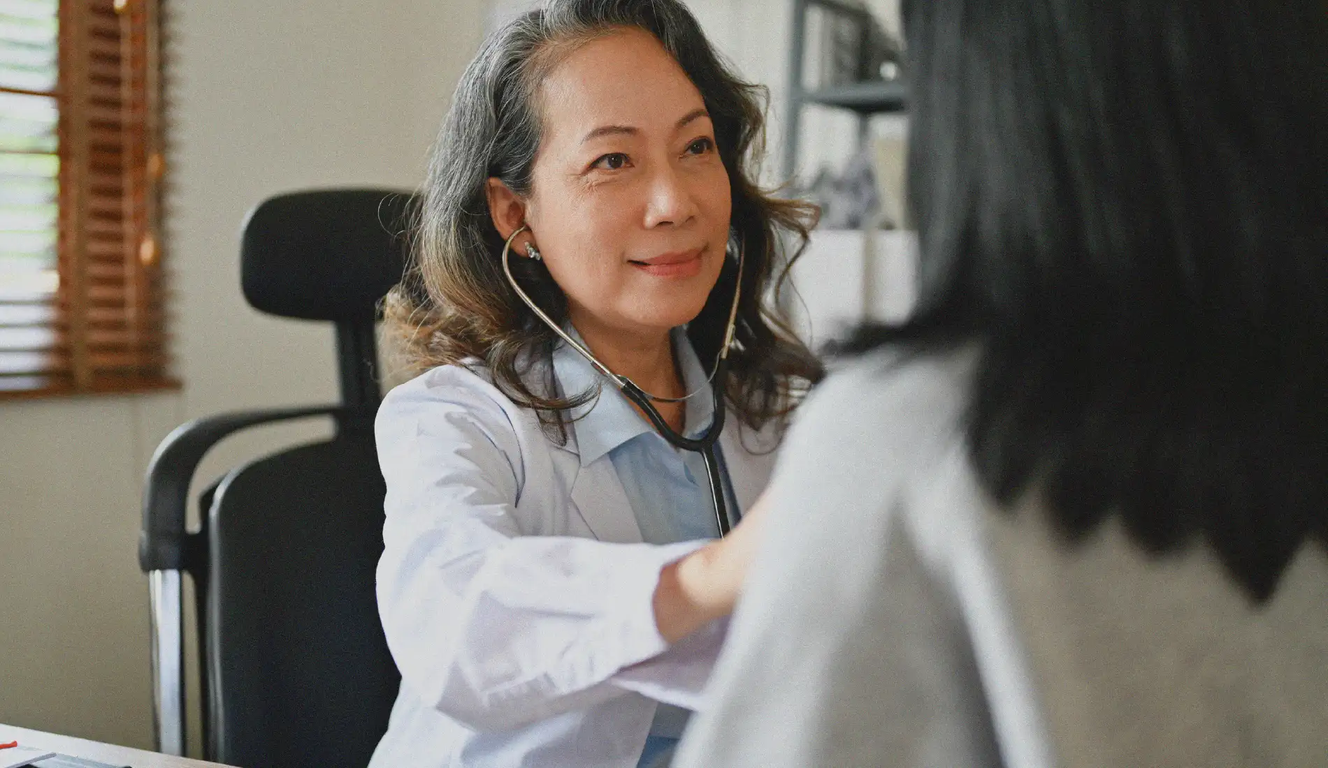 Une femme médecin qui ausculte une autre femme qui a le dos tourné à l'objectif. On ne voit que ses cheveux noirs qui retombent sur ses épaules.