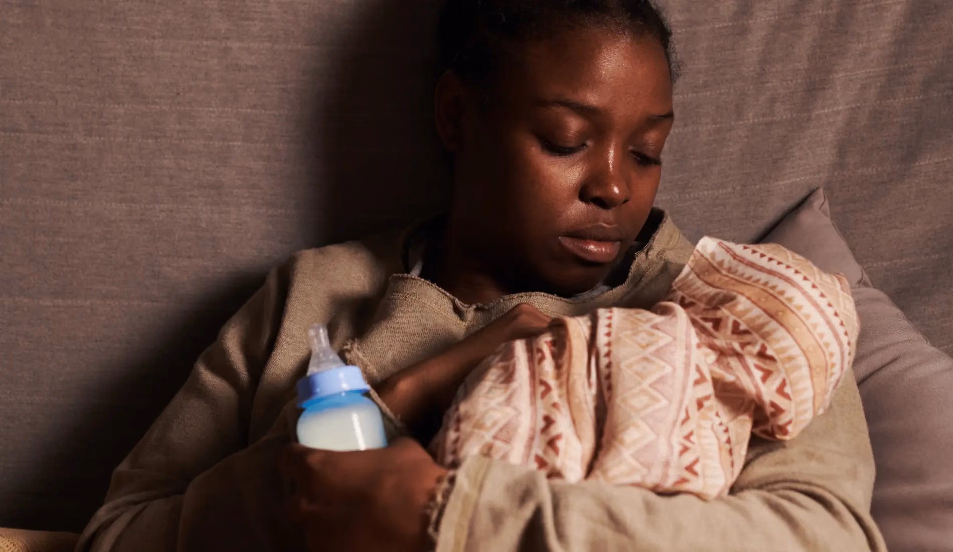 Jeune mère épuisée affalée dans un canapé, bébé dans les bras et biberon à la main, illustrant les effets de la charge mentale des mères sur la santé et le sommeil.
