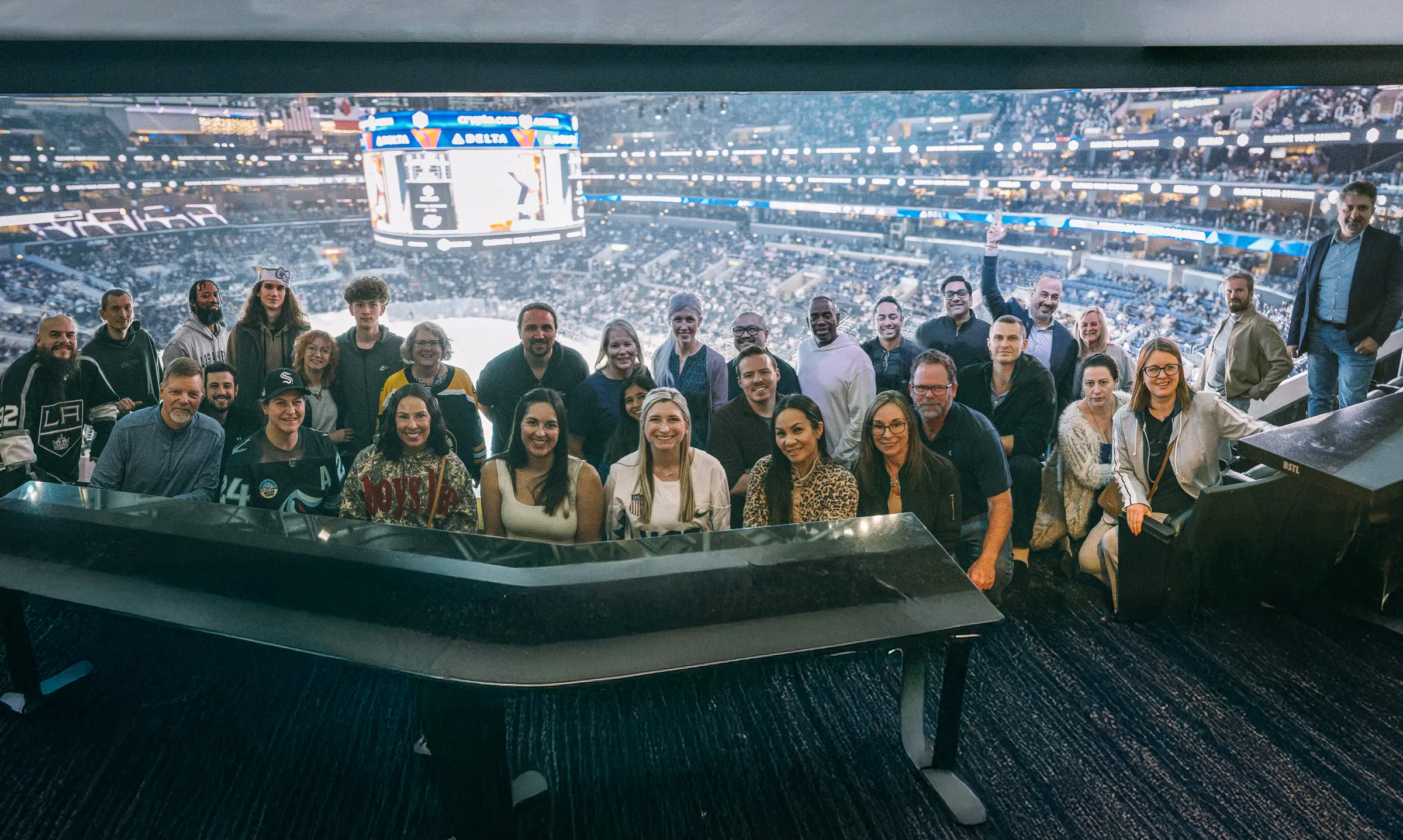 A large group of people posing together in a private suite at a hockey arena, smiling in front of the rink and scoreboard with the crowd filling the stadium behind them.