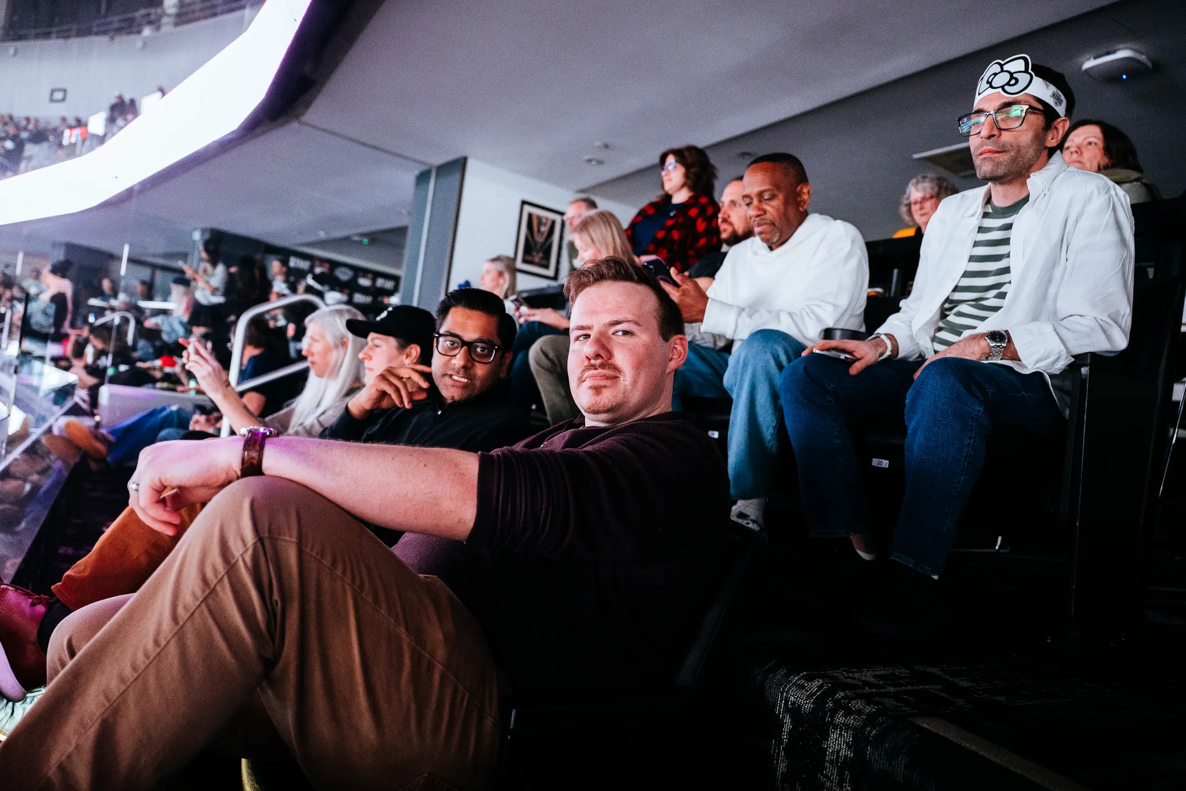 A group of people sitting in arena seats at a hockey game, with one man in the foreground looking at the camera while others watch the action or check their phones.