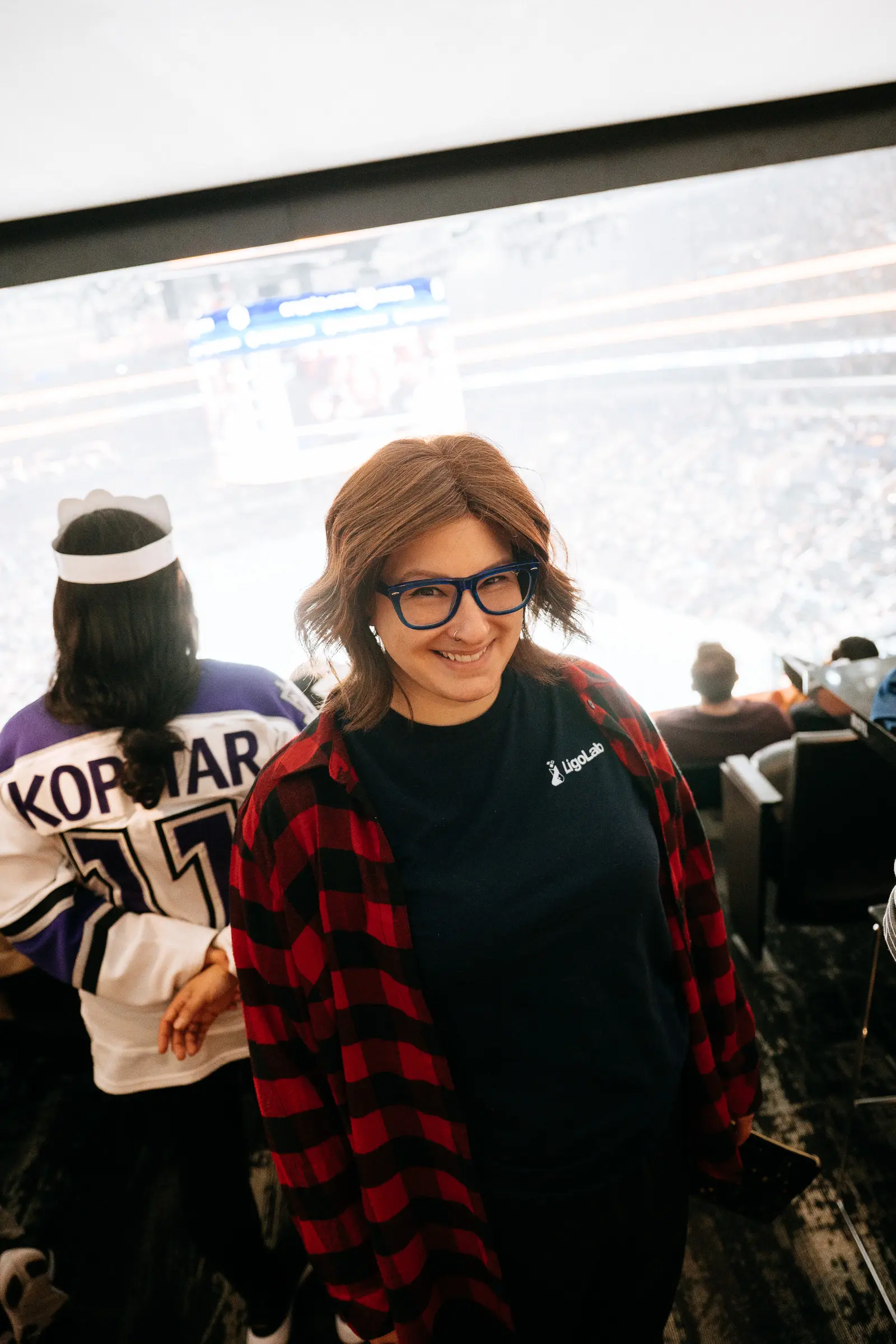 A smiling woman wearing blue glasses, a black shirt, and a red plaid flannel stands in arena seating at a hockey game, with the rink and crowd visible in the background.
